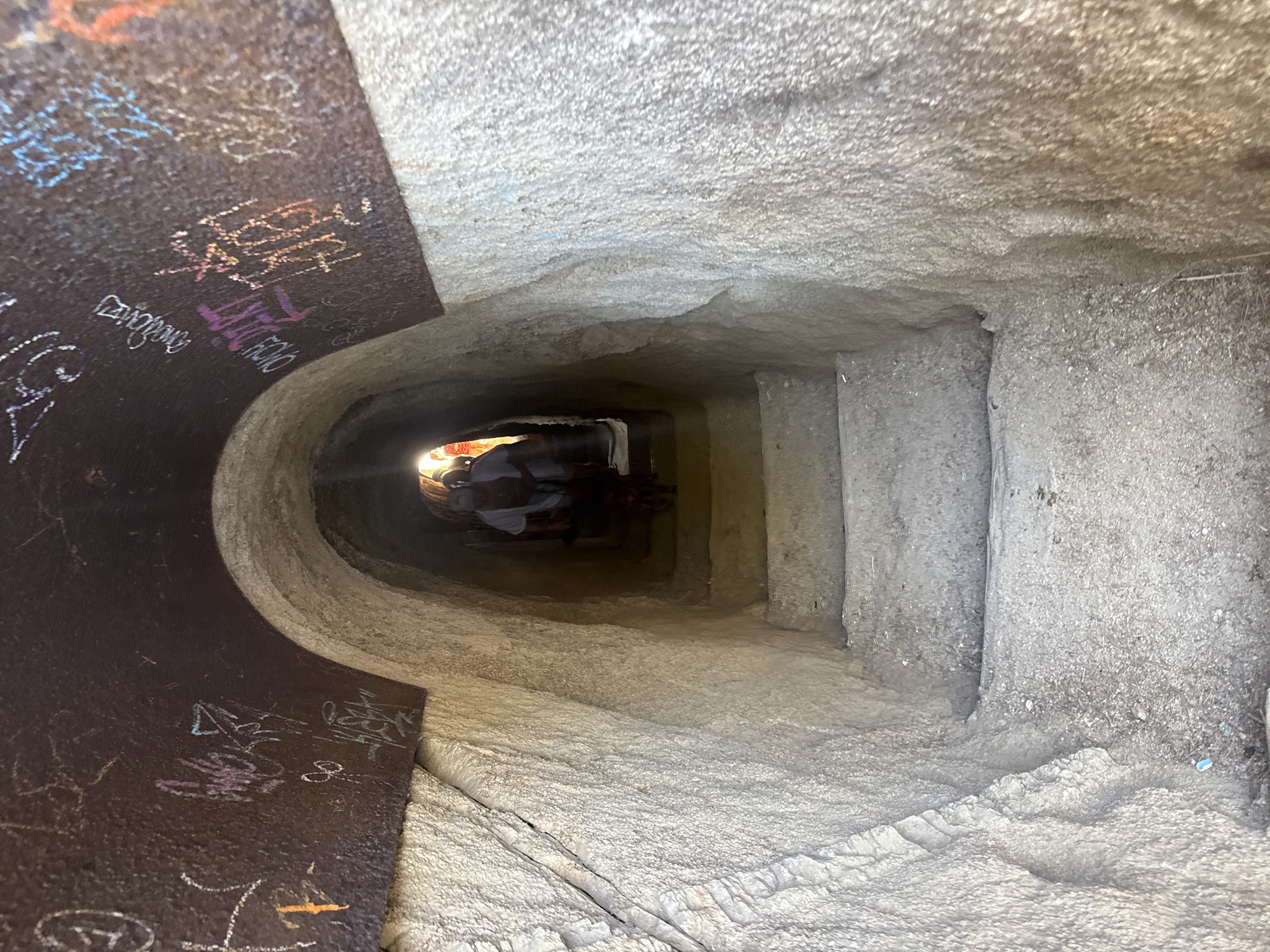 The image depicts the entrance to a tunnel or passage carved into rock, likely a historical or man-made structure. The tunnel has a rough, natural stone interior and is partially covered by a metal ceiling that is adorned with graffiti in various colors, including purple, pink, orange, blue, and white. The graffiti includes stylized lettering and symbols.
A set of rough-hewn stone stairs descends into the dimly lit interior of the tunnel. At the bottom of the visible stairs, a person wearing a light-colored shirt is partially visible, walking further into the tunnel. A small animal, possibly a dog, is walking in front of the person. The light at the end of the tunnel suggests an opening or exit. The overall impression is of an underground passage within a rocky landscape.
This scene is located in Castell d'Aro, Platja d'Aro i s'Agaró, Spain, likely within a historical site or natural attraction that has been adapted for access.