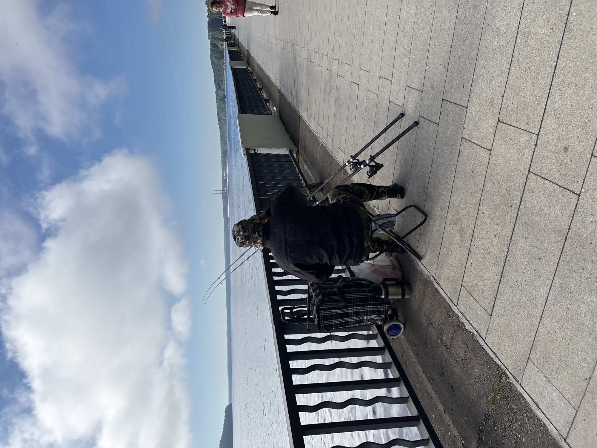 The image shows a man fishing on a waterfront promenade in Chaikovsky, Russia. The man is seated on a folding chair, wearing a camouflage hat and jacket, and camouflage pants. He is facing away from the camera, towards the body of water. Two fishing rods are set up in rod holders in front of him. Beside him is a checkered shopping cart with a thermos and a plastic bag.
The promenade is paved with light-colored stone tiles. A black metal railing separates the promenade from the water, which is calm and reflects the blue sky and scattered white clouds. In the distance, along the far shore of the water, a line of trees and buildings is visible, including two prominent industrial smokestacks.
To the right of the frame, a woman is walking along the promenade. She is wearing a light-colored hooded jacket and white pants. The scene appears to be during the daytime, with bright sunlight. The overall environment suggests a recreational area by a large body of water.