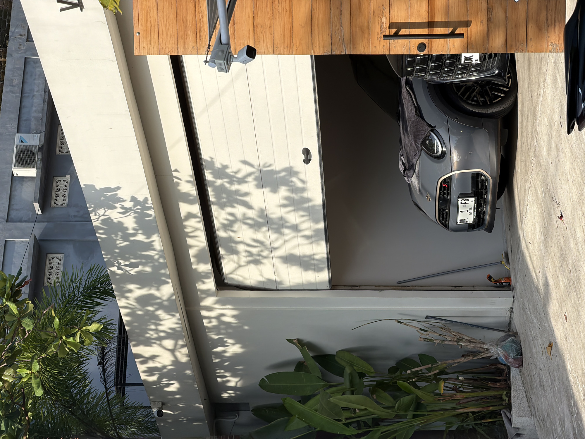 An open garage door reveals a gray car parked inside a garage on a sunny day. Outside, tropical plants grow near the building, and tree shadows are cast on the structure. The image appears to be taken in Mengwi, Indonesia.