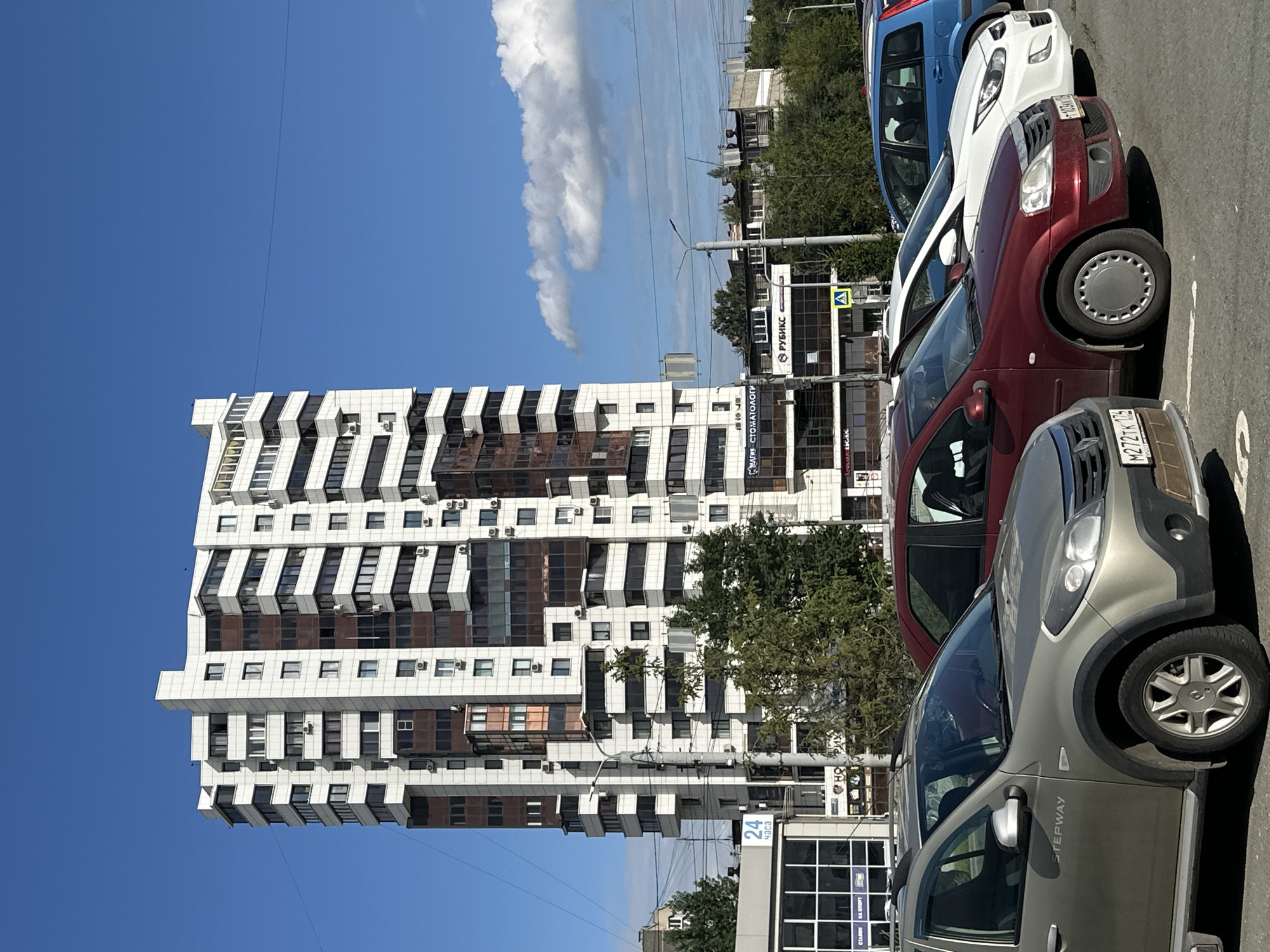 A multi-story, modern apartment building with white and brown facade dominates the background of this outdoor daytime scene. The building features numerous balconies and windows, with air conditioning units visible on the exterior. In the foreground, a row of parked cars is visible, including a silver SUV, a red sedan, a white sedan, and a blue minivan. The vehicles are parked in designated spots on an asphalt surface. The environment appears to be an urban setting, with trees and other buildings visible in the midground. The sky is blue with scattered white clouds.