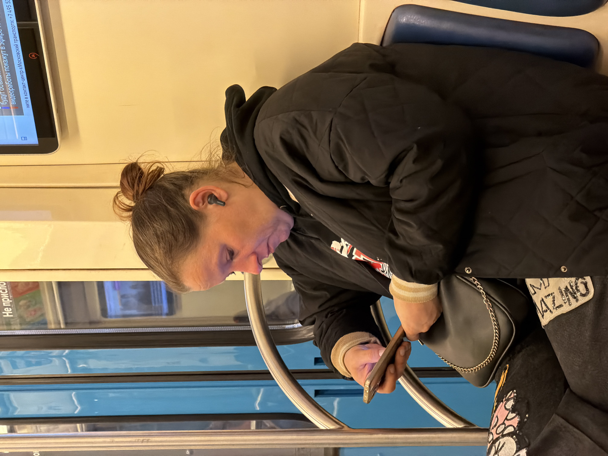 The image shows a woman sitting on what appears to be public transportation in Moscow, Russia, based on the Cyrillic text displayed on the nearby monitor. The woman has her hair in a bun and is wearing an earbud in her left ear. She is dressed in a black puffer jacket over a hoodie, and her pants have decorative elements. She is holding a smartphone in her hands and appears to be using it. A small black purse with a chain strap rests beside her. The interior of the transportation vehicle is visible, with blue panels and metal handrails.