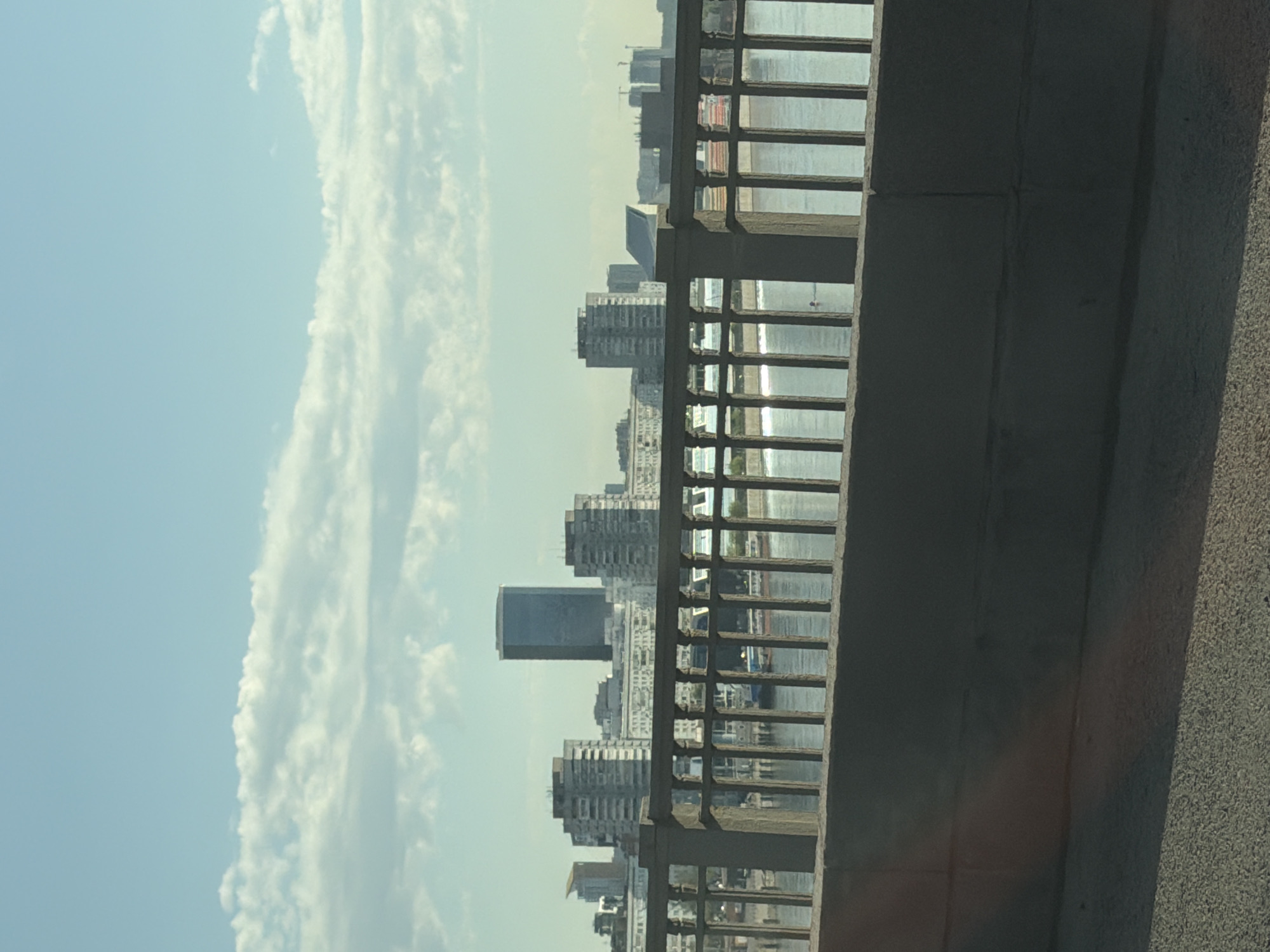 The image shows a cityscape viewed through the railing of a bridge. The sky is light blue with white clouds. Multiple tall buildings are visible in the distance, including a dark, rectangular skyscraper. Water, likely a river, is seen between the railing and the buildings. The bridge railing is made of concrete and metal bars. The location is likely Krasnoyarsk, Russia, given the context. The scene appears to be taken on a sunny day. No people are clearly visible.