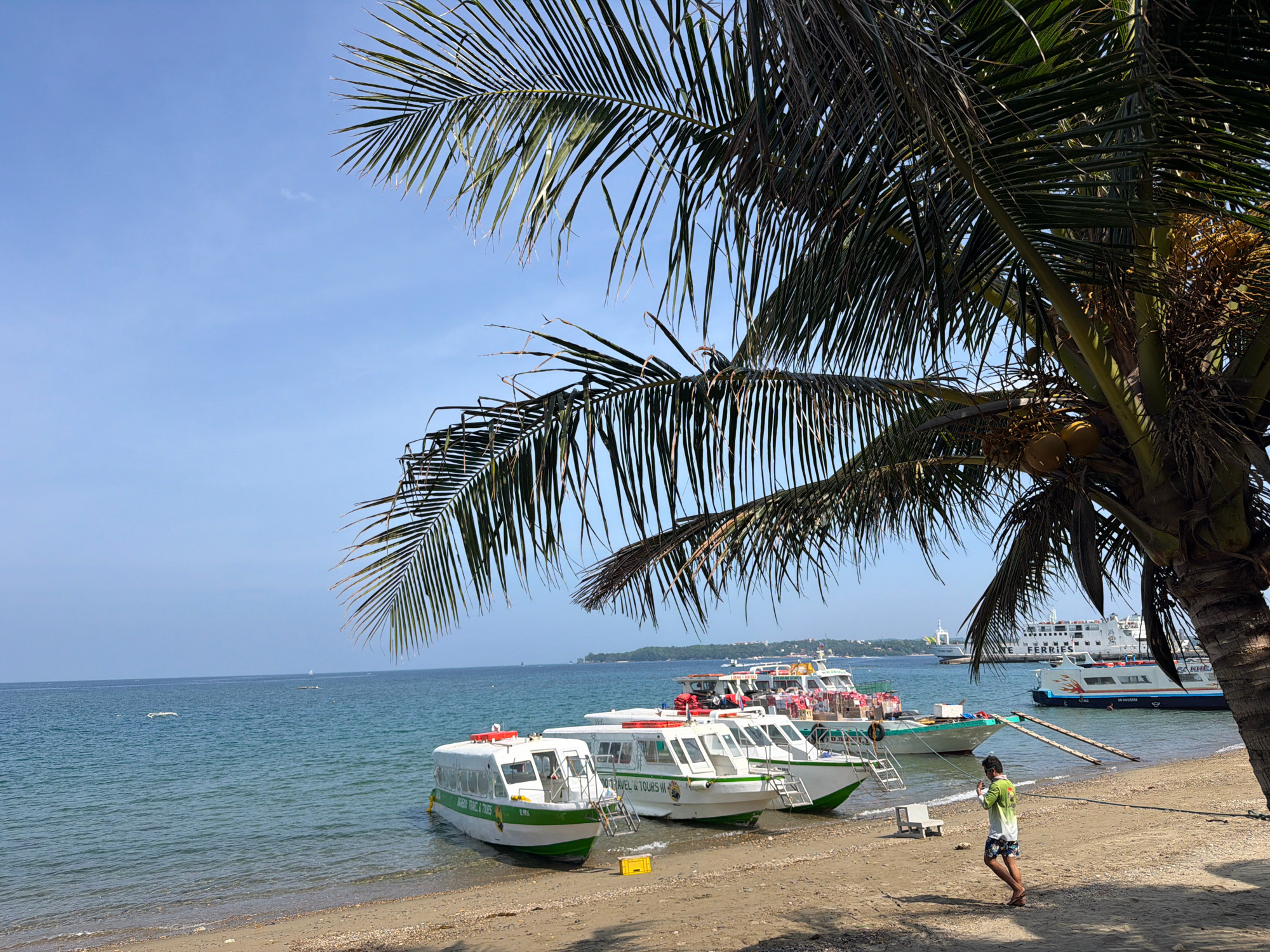 In Malay, Philippines, a tropical beach scene unfolds under a clear blue sky, fringed by the lush fronds of a palm tree in the foreground. Several white passenger boats, some adorned with green trim, are moored close to the shore. One boat, labeled "BAGUIO TRAVEL & TOURS III," has its name clearly visible. A lone man, wearing a light green t-shirt and patterned shorts, walks along the sandy beach, his back partially to the viewer. In the background, larger ferries are anchored in the turquoise water, suggesting a bustling port or transportation hub. The beach itself is a mix of sand and small pebbles, with a few scattered items including a yellow container and a small white bench.
