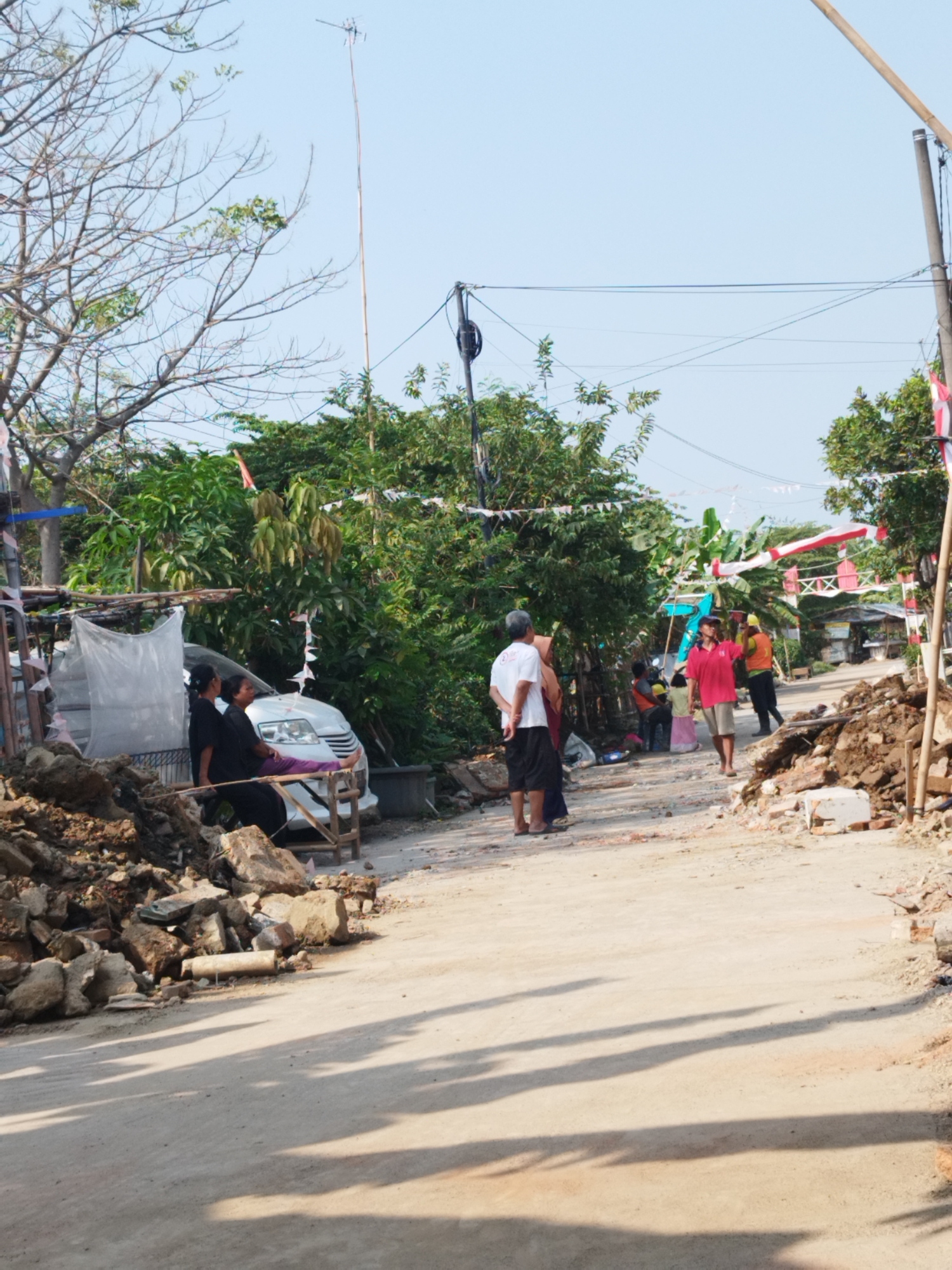 In Karawang, Indonesia, a dirt road is visible, lined with piles of rubble on either side, suggesting construction or demolition. Overhead, power lines crisscross the sky, strung between utility poles. Several people are present. On the left, two women are seated on a wooden structure next to a white car. Further down the road, a group of people are gathered, with one person in a red shirt and khaki shorts standing in the center, appearing to speak or gesture. Another person in a yellow vest and hard hat is visible behind him. Red and white bunting, likely Indonesian flags, are strung between trees and poles, decorating the area. The environment appears somewhat rustic, with lush greenery bordering the road.