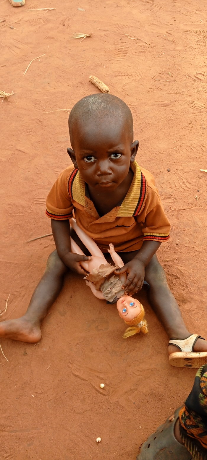 A dark-skinned child is seated directly on reddish-brown earth in Ngalula, Democratic Republic of the Congo. The child, identifiable as a young boy, wears an orange short-sleeved polo shirt featuring yellow and red striped accents on the collar and sleeve cuffs, and a small dark emblem on the left chest. He has short, dark hair and looks directly forward with a somber expression. His left foot is bare, while his right foot is partially covered by a light-colored sandal. The child holds a fair-skinned doll with blue eyes and blonde hair styled in pigtails; the doll is dressed in a tattered, light-brown garment. The ground surface is uniformly reddish-brown, exhibiting multiple faint footprints, scattered fragments of dried vegetation, and a single corn cob piece situated behind the child's head. Two small, light-colored seeds or pellets are visible on the ground near the child's right leg. A portion of a second individual's leg, clad in dark footwear and patterned fabric, is visible in the lower right corner of the frame.
