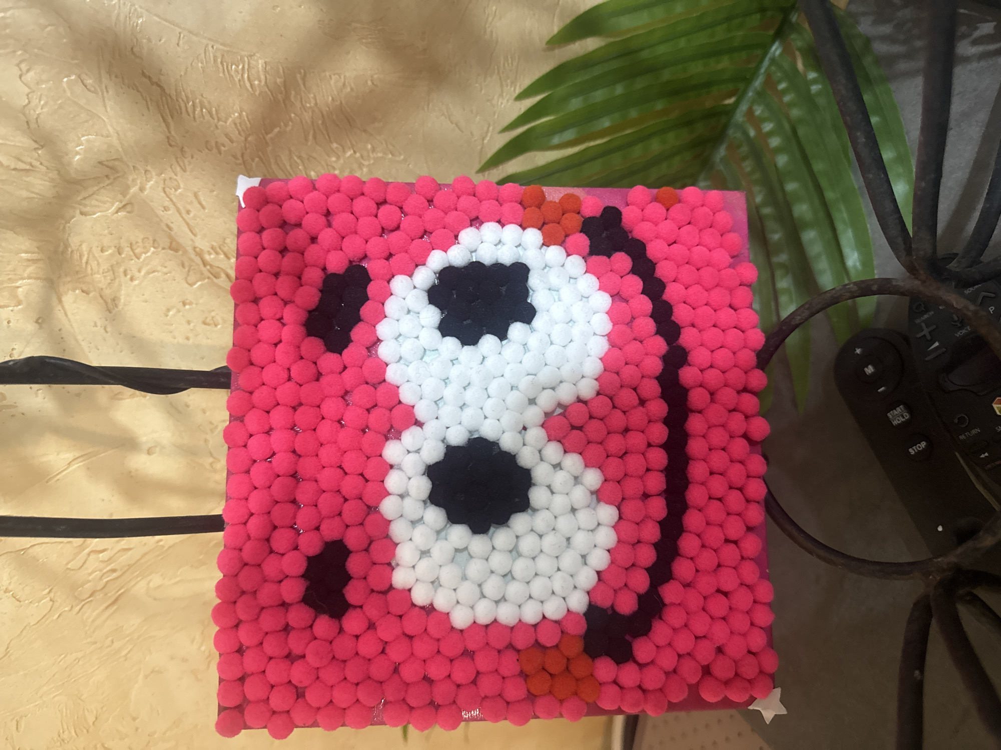 A square pink object is decorated with pink, white, black, and orange pom-poms in a design that resembles a panda bear holding a soccer ball. The object sits on a surface and is partially obscured by black metal bars on the left and right. A green plant leaf is visible in the background. A black remote control sits to the right of the square pink object. The exact location is undetermined.