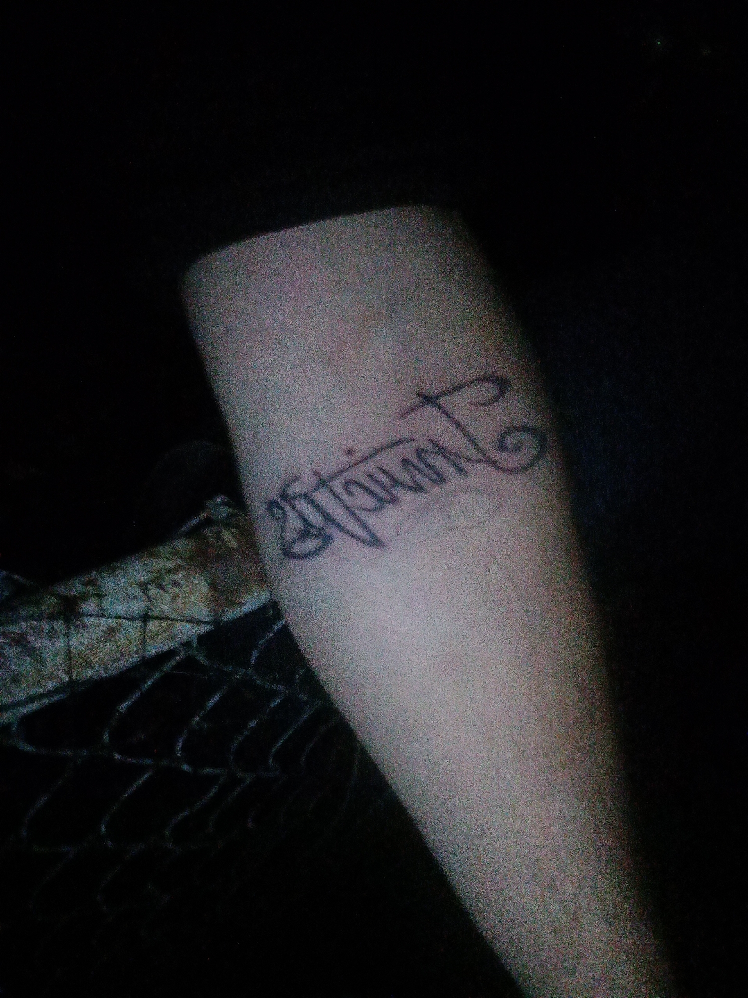 The image shows a tattoo on a person's arm, with a dark background. The tattoo is a word written in a cursive script, potentially in an Indian language, though specific identification is difficult due to image quality. The surrounding environment is dark and unclear, with what appears to be a fence partially visible. The location in Tahlequah, United States of America is not known from this image.