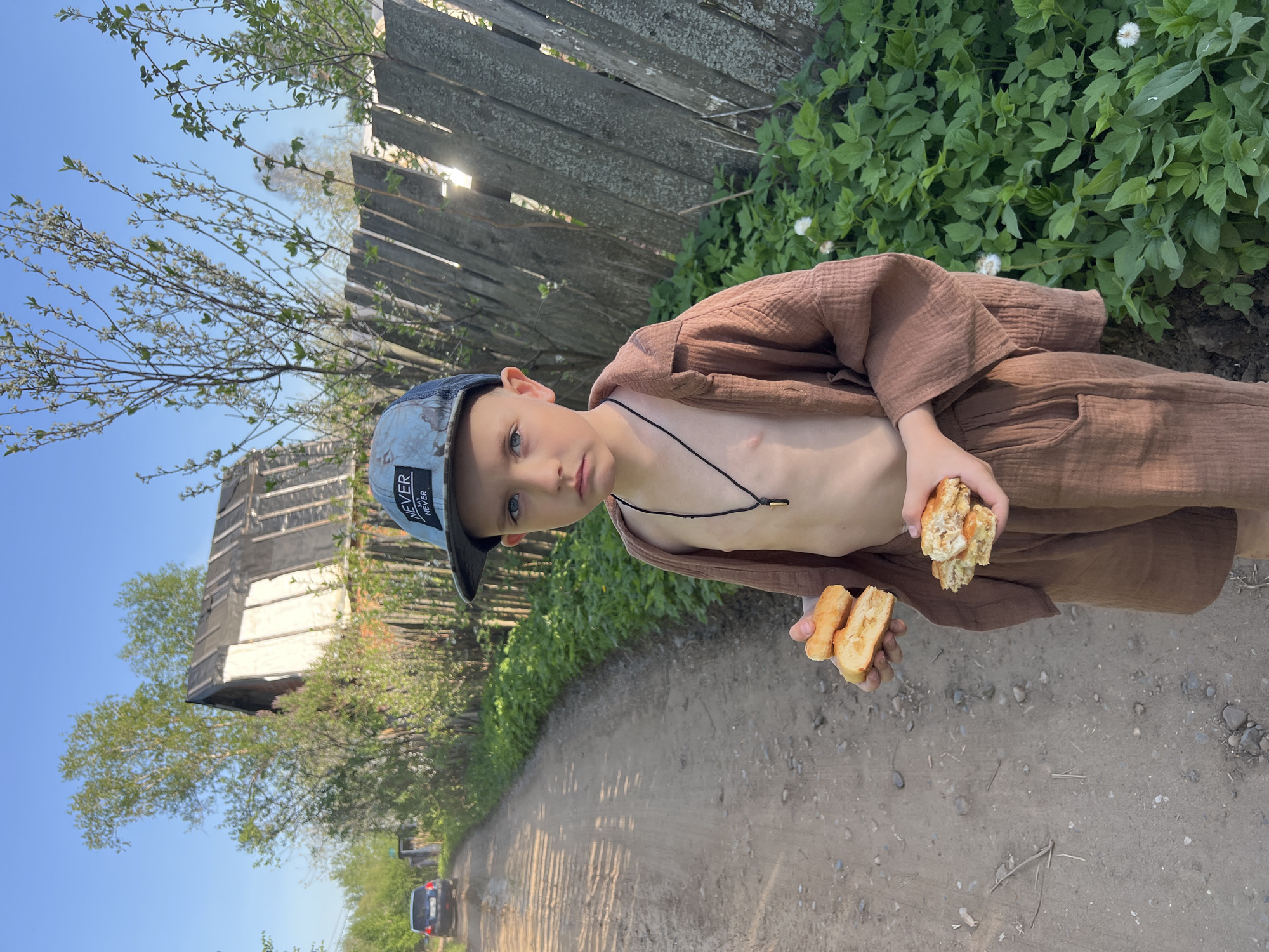 A young boy stands on a dirt road next to a wooden fence, dressed in a brown shirt and pants, wearing a cap with the word "NEVER" on it. He holds bread in both hands. In the background are green plants, a dilapidated structure, a road with a car in the distance, and a blue sky. The location is likely in a rural or semi-rural area near Noviy, Russia, based on the architecture and environment.