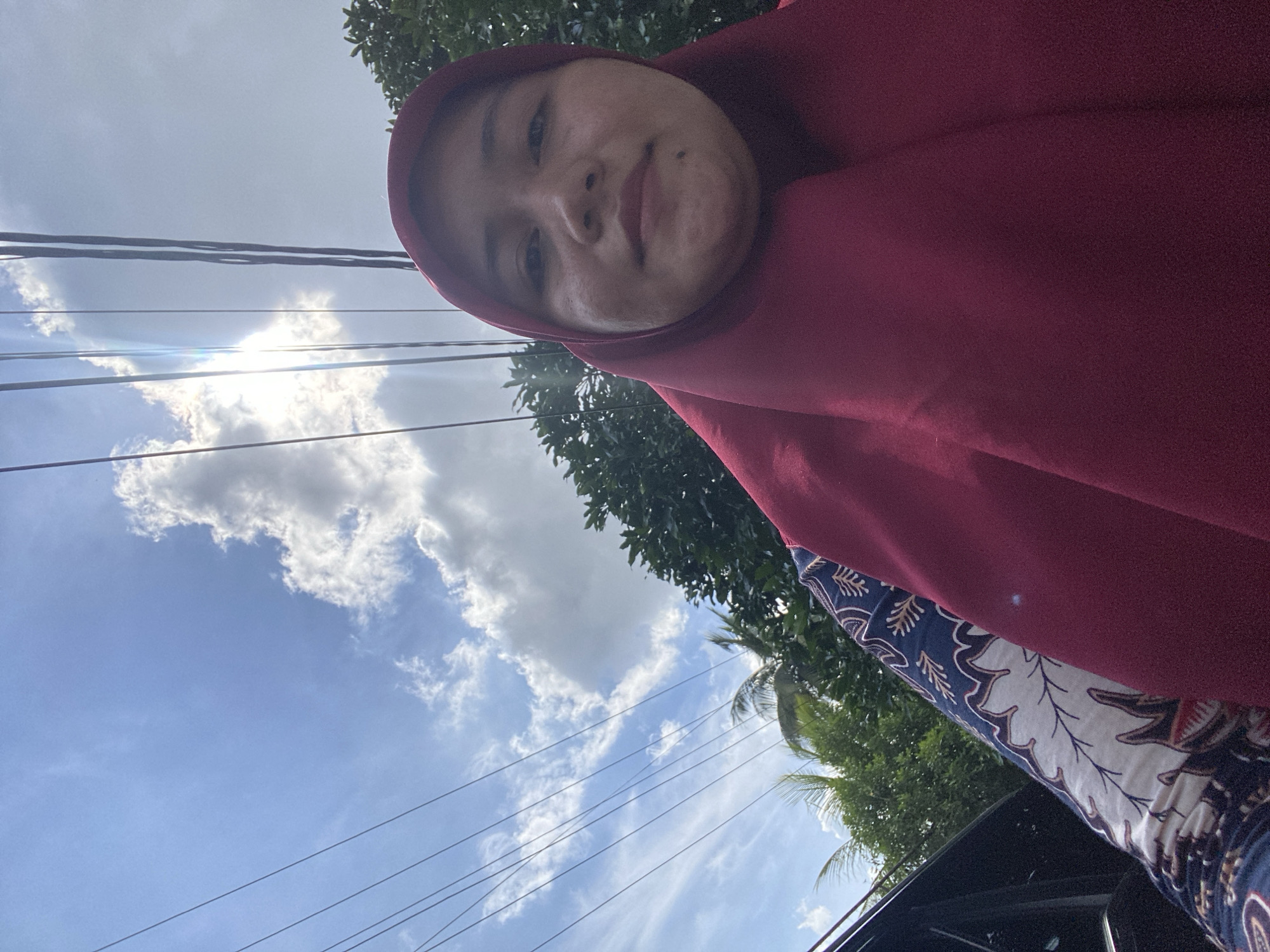 A selfie is taken from a low angle, capturing a woman wearing a maroon hijab and a batik-patterned garment. She is outdoors, with a bright blue sky filled with scattered clouds visible behind her. Sunlight beams through the clouds, creating a lens flare effect. Several power lines run diagonally across the sky. Lush green trees are present in the background, and a portion of a dark-colored car is visible on the left side of the frame. The woman's face is turned towards the camera, and she has a mole on her chin. The setting appears to be a residential or semi-rural area within Banjarbaru, Indonesia, suggested by the combination of overcast sky, power lines, and abundant greenery.