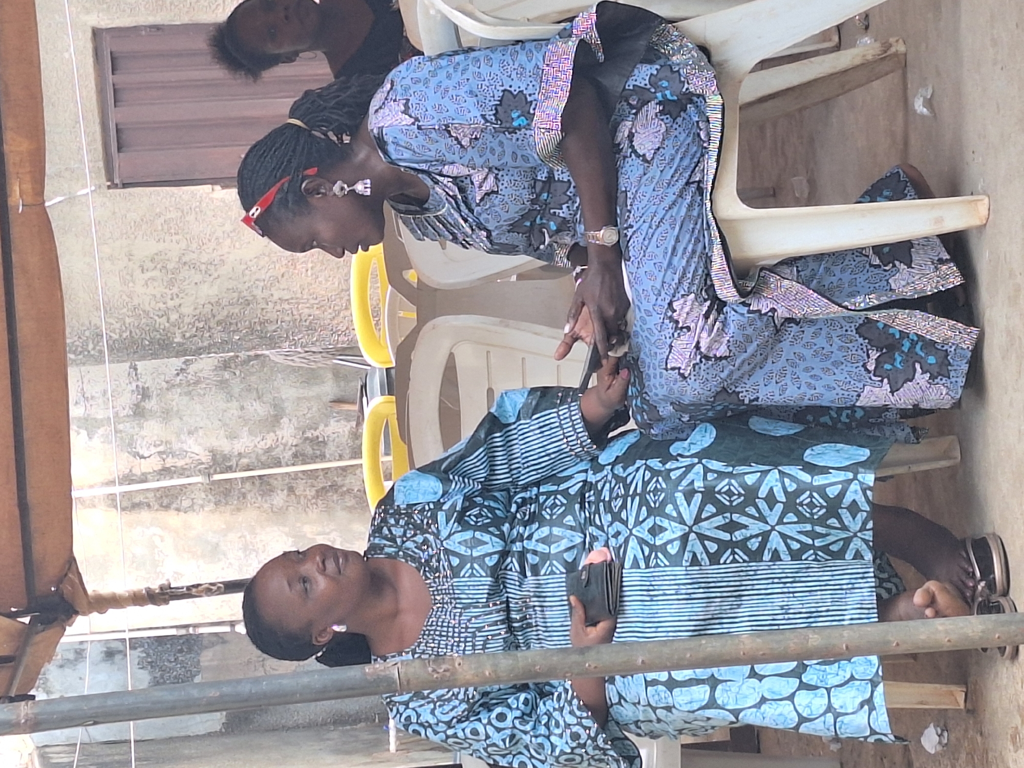 Two women are seated on plastic chairs in Sabon Tasha, Nigeria. The woman on the left is holding a black wallet or phone. Both women are wearing similar patterned dresses, with the seated woman above bending forward. A younger person is partly visible in the background. The area appears to be an outdoor space near a building. Metal piping is in the foreground.