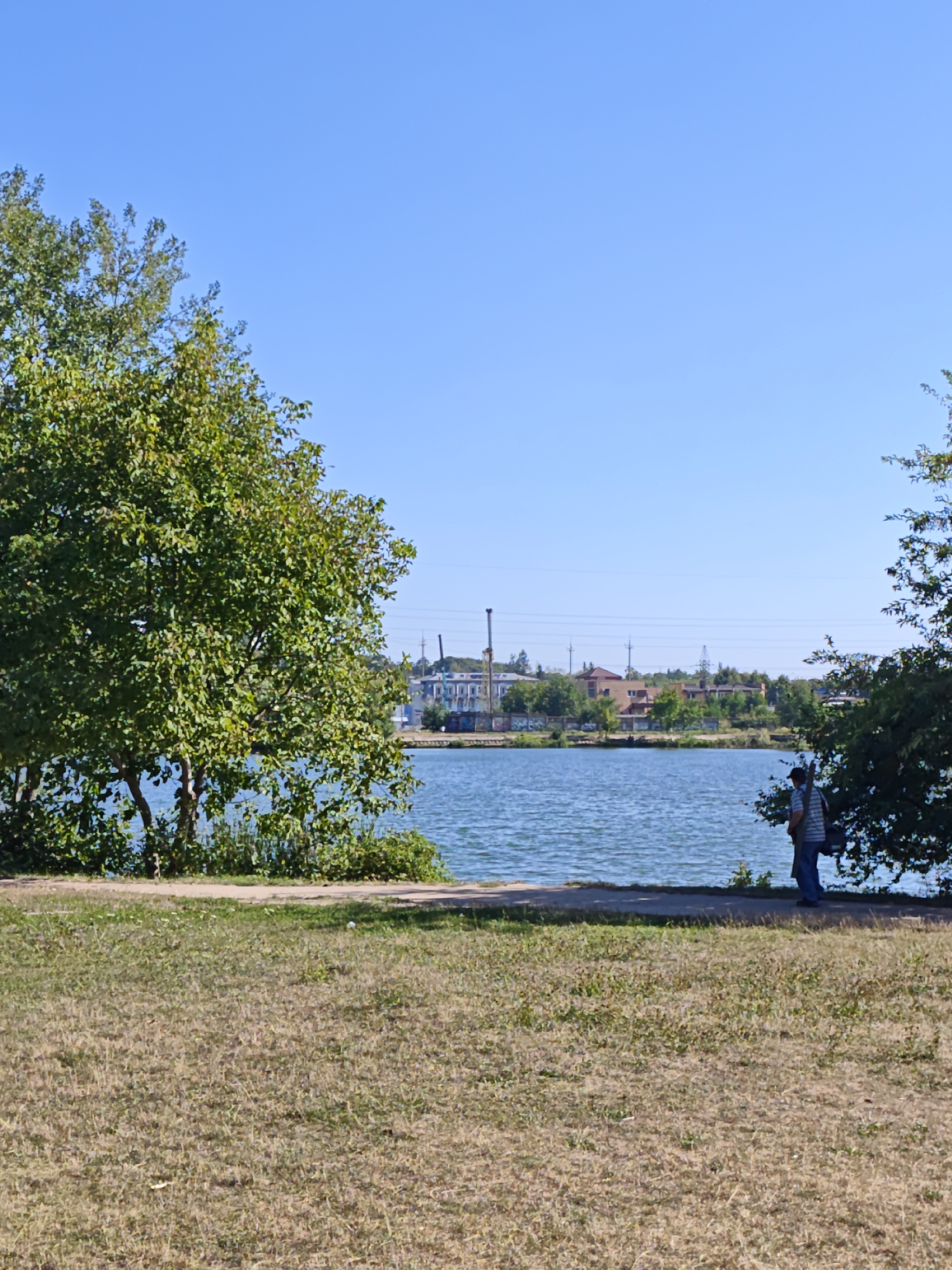 This is a vertically oriented, eye-level shot of a waterfront scene under a clear blue sky. In the foreground, dry, brown grass covers the ground. A paved path runs horizontally across the middle of the image, separating the grassy area from the water. The water of a lake or river is a calm, blue expanse. On the far side of the water, a treeline with green foliage is visible, interspersed with buildings and industrial structures, including chimneys and power lines. On the right side of the image, a person wearing a striped shirt and blue jeans stands near the water, facing away from the camera and towards the water and distant buildings. Trees with green leaves frame the right side of the image. On the left side, a large tree with lush green foliage dominates the foreground, partially obscuring the view of the water and the distant shore. The scene appears to be a public park or recreational area in Vinnytsia, Ukraine, likely along the Southern Bug river.