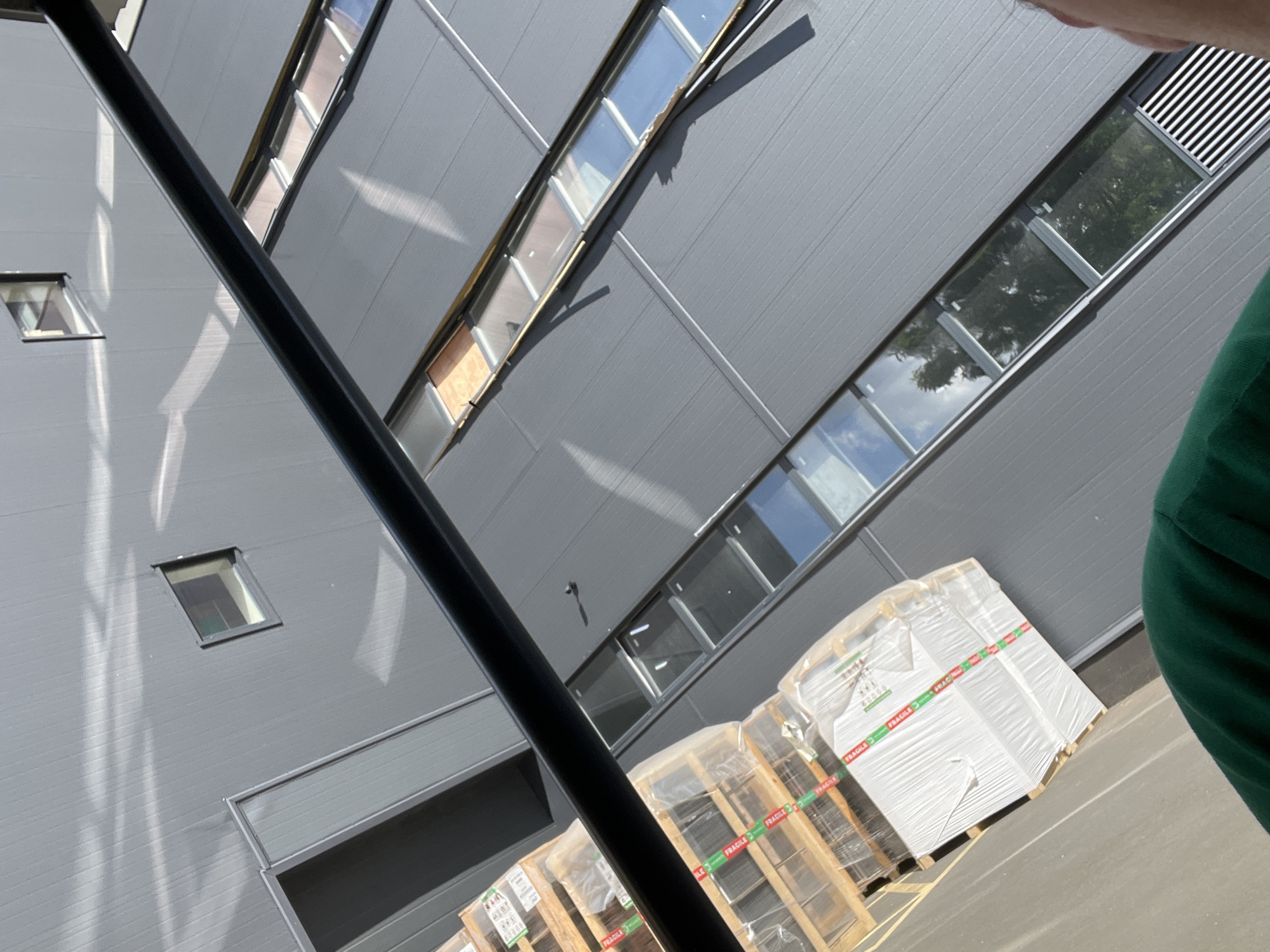 The image shows a close-up, angled view of the exterior of a modern, grey industrial building, likely a warehouse or distribution center. The building has a corrugated metal facade and a series of long, narrow windows set high up, reflecting the sky and surrounding trees. In the foreground, several pallets of goods wrapped in plastic are visible, some with "FRAGILE" tape. A dark, horizontal beam runs across the top of the frame, obscuring a portion of the building's upper section. The lighting suggests a sunny day, with strong shadows cast on the building's surface. The scene appears to be outdoors, possibly in an industrial zone or logistics hub. There is no direct indication of the city being Kyiv, Ukraine, but the subject matter and architecture are consistent with such a location. A small portion of a person's shoulder and arm, wearing a green garment, is visible in the bottom left corner of the image.