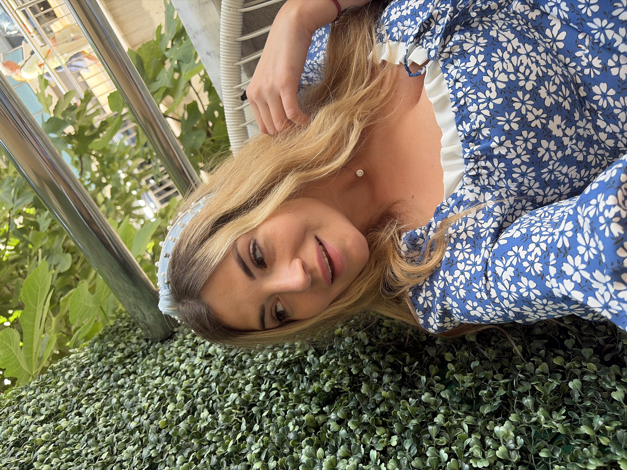 A medium shot photograph captures a fair-skinned woman with blonde hair and light brown eyes, looking to her left. She is wearing a blue dress with a white floral pattern and a pearl-embellished headband. She is seated in a white wicker chair, with a lush green hedge wall to her left and a metal railing and foliage in the background. In the blurred background, two individuals can be seen standing on what appears to be a balcony or promenade. The scene is set outdoors, with natural light suggesting daytime. The overall impression is casual and relaxed. Based on the visual elements, this scene is likely located in Budva, Montenegro, a popular coastal city known for its vibrant atmosphere and scenic views.