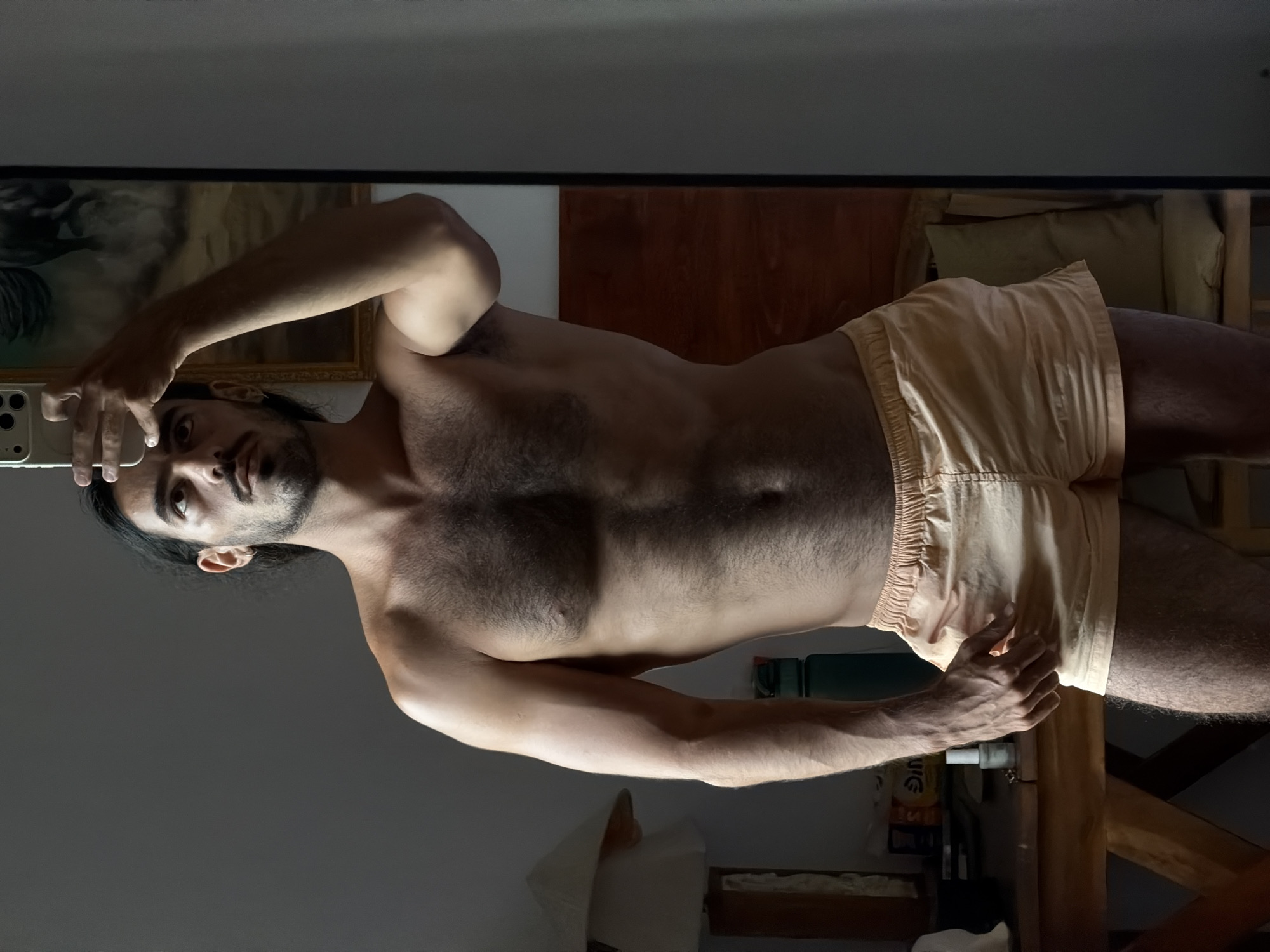 A man is photographed in a mirror selfie. He is shirtless, wearing tan shorts, and appears to be in a domestic setting, with furniture and objects visible in the background. There are no details to indicate the location is North Kuta, Indonesia.