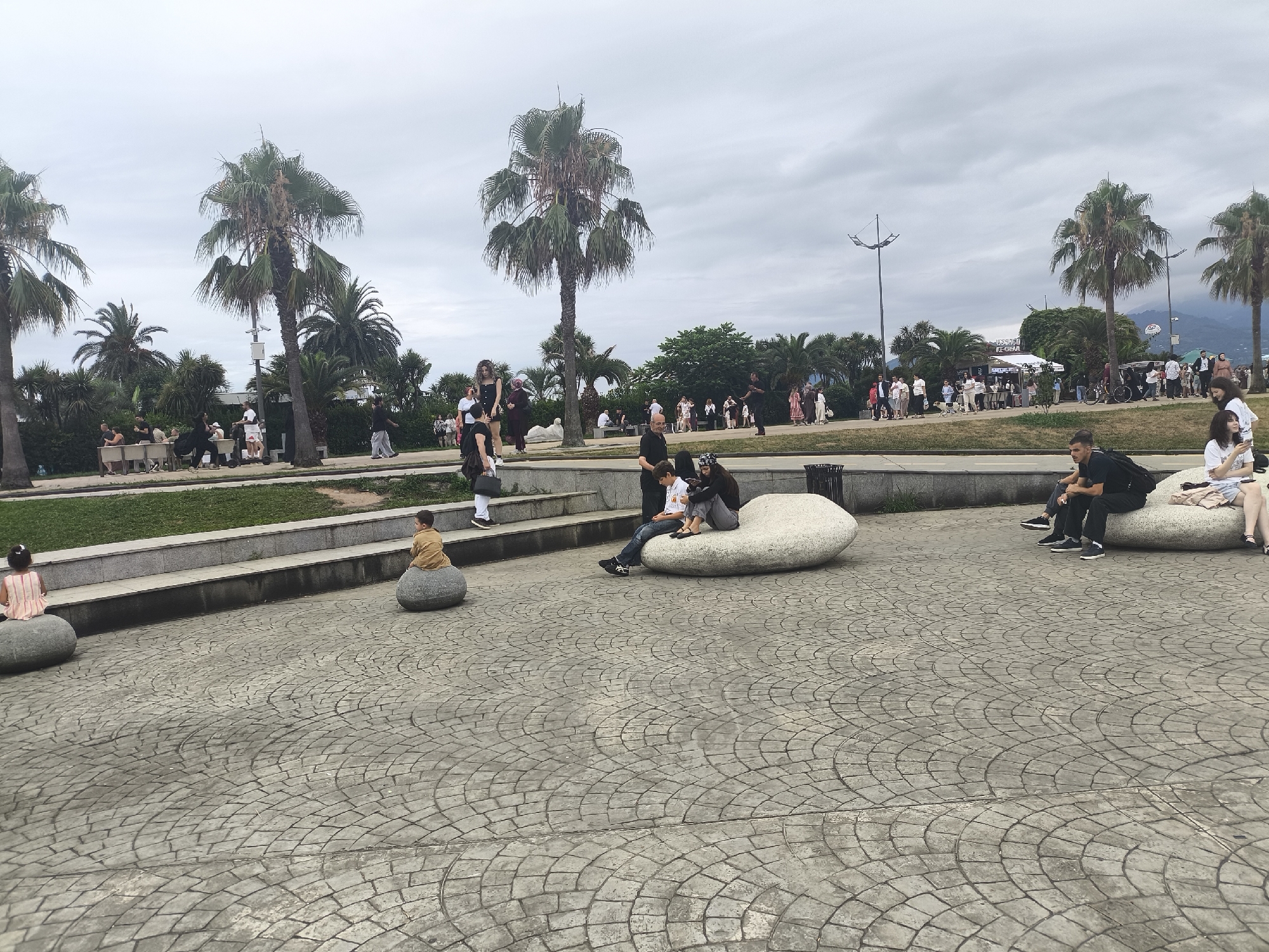 This is an eye-level, medium shot of a paved plaza in Batumi, Georgia, likely near the waterfront. The plaza is made of grey cobblestones with a repeating fan pattern. Two children are seated on large, smooth, light-colored boulders in the foreground. Behind them, a group of people are seated on similar boulders or benches in a shaded area beneath palm trees. Further in the background, more people are walking and standing, and a line of food stalls with awnings are visible. The sky is overcast and grey. Palm trees are scattered throughout the scene, with some taller ones in the middle ground and a cluster of shorter ones to the left. In the far background, mountains are visible through the haze.