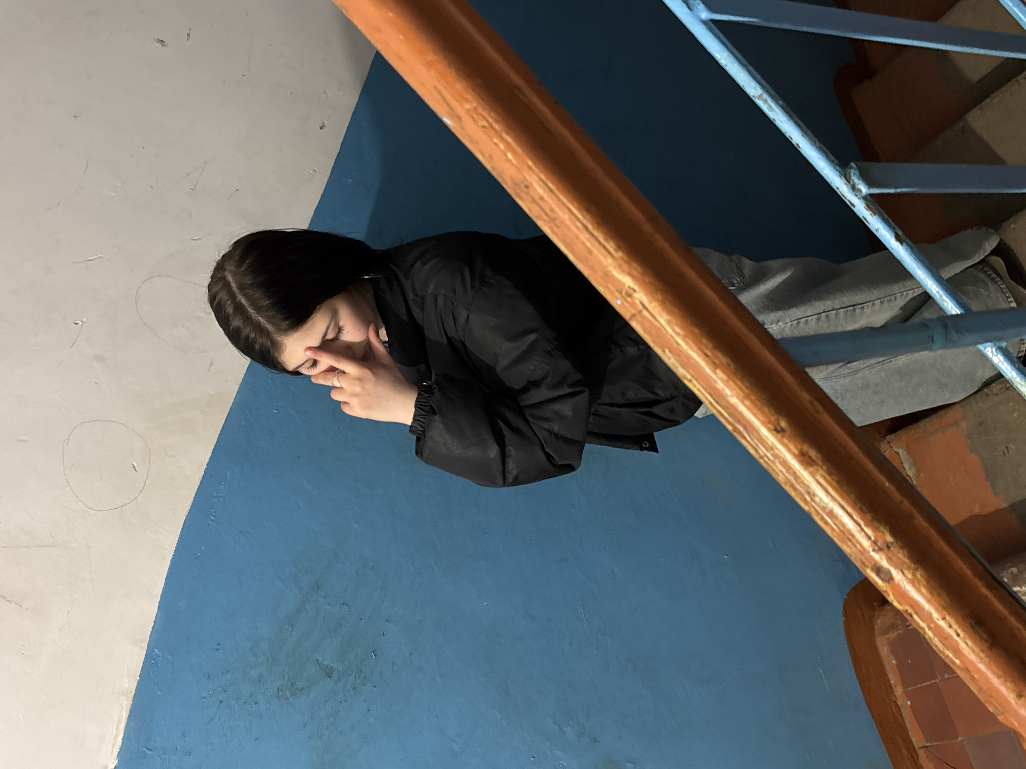 A young woman with dark hair is depicted lying or sitting on the blue-painted floor of a building's interior, likely a stairwell landing. She is dressed in a black jacket and light-colored jeans. Her right hand covers her mouth and part of her nose, her eyes are closed or looking downwards, conveying an emotion of sadness, distress, or exhaustion.
To her right, a sturdy wooden handrail runs diagonally upwards, part of a staircase structure. Below this handrail, a light blue metal railing is visible, behind which brown-orange tiled steps can be seen. The wall adjacent to where the woman is positioned is painted in two sections: a vibrant blue on the lower half (matching the floor) and a lighter white on the upper half, meeting at a curved line. The overall environment, with its painted walls and worn railings, suggests an older, possibly public or residential building. The lighting is somewhat dim, implying an indoor setting possibly illuminated by artificial light. There is no visible text in the image.