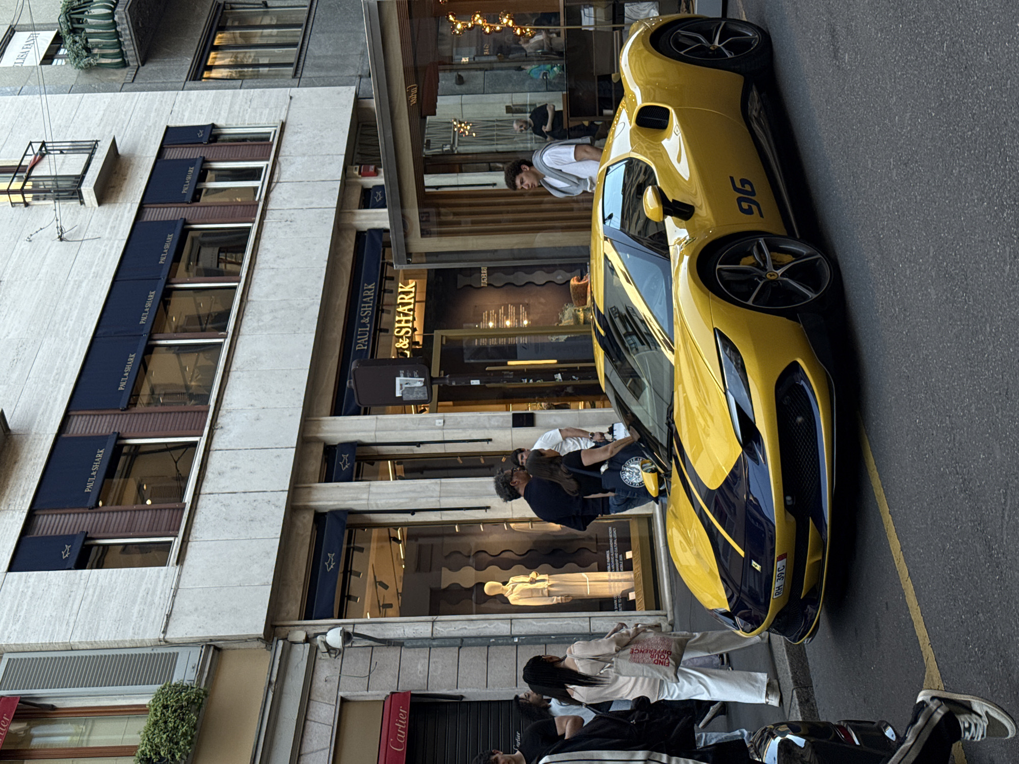 The image shows a vibrant yellow sports car parked on a street in Milan, Italy, adjacent to a sidewalk filled with pedestrians. The car has a prominent "96" printed on its side. On the left side of the frame, a multi-story building houses retail stores, including "Paul & Shark" and "Cartier," identified by their signage. Several people are standing or walking on the sidewalk, with one group appearing to interact near the car. The overall scene conveys a bustling urban environment with a mix of luxury retail and pedestrian traffic.