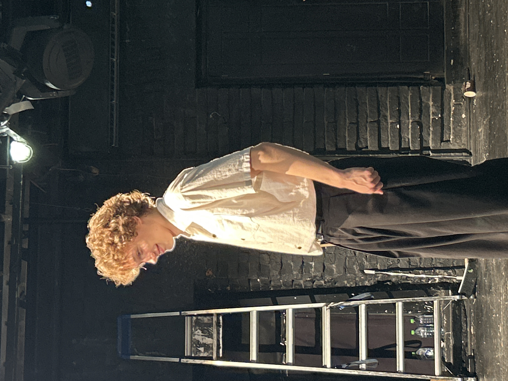 A young man with curly, light brown hair stands on a stage, looking down and to his left with a slight smile. He is wearing a short-sleeved, cream-colored button-up shirt with the top two buttons undone, and loose-fitting black pants. His right arm is bent at the elbow, with his hand resting near his hip.
To the left of the man is a silver stepladder with blue accents. Several bottles of water with green caps are visible on one of the lower steps. A broom leans against a dark brick wall behind the ladder. The wall extends to the right and is partially obscured by the man. A closed, dark door is visible on the far right side of the frame. Overhead, stage lighting fixtures are present. The floor appears to be wet and reflective, with some debris scattered on it, including a small, discarded paper cup. The background is dark, suggesting an indoor performance space or studio.