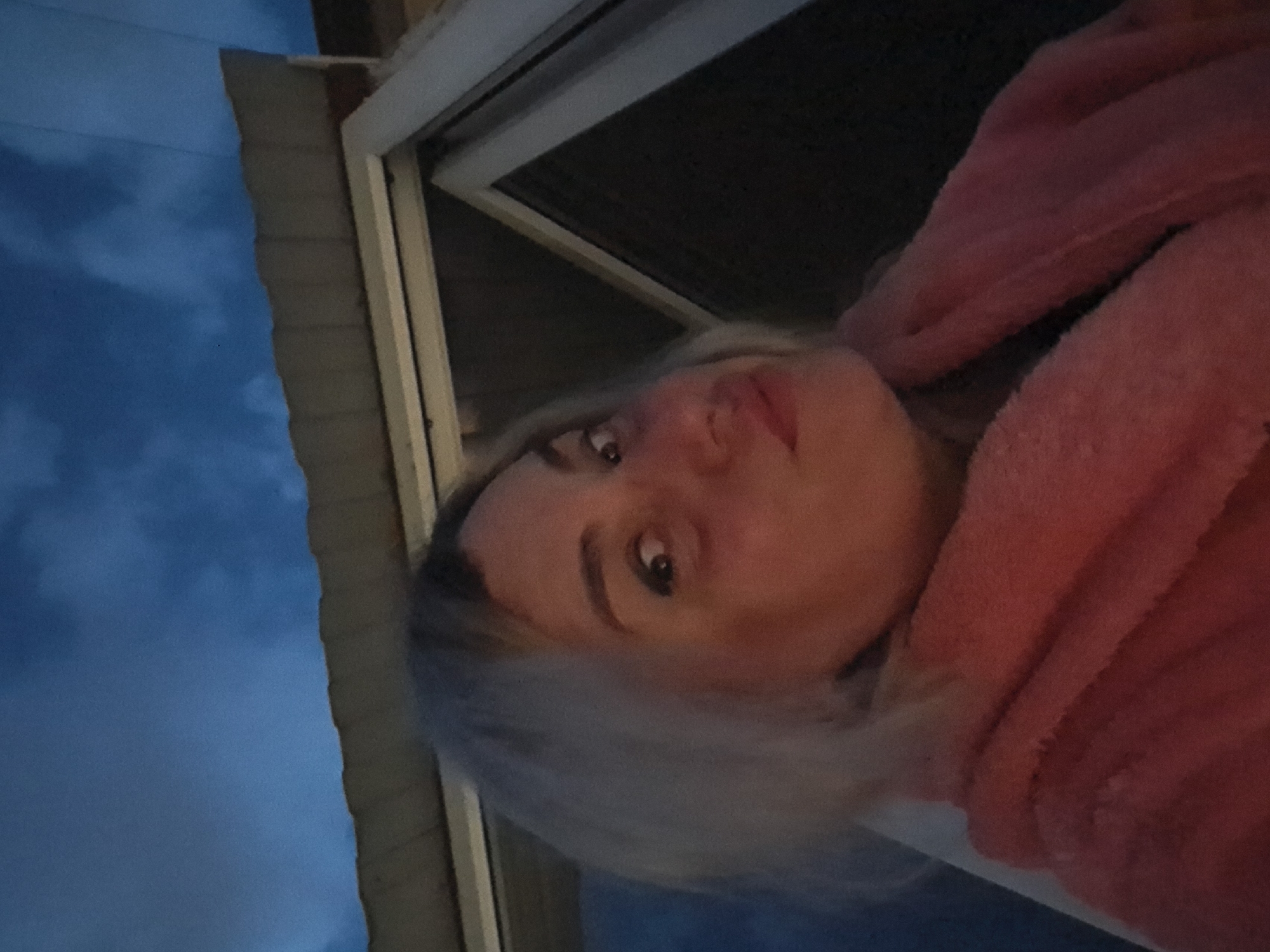 A woman with blond hair and wearing a pink fuzzy robe is visible inside a building, possibly leaning out a window. Outside the window, a section of a blue, cloudy sky is visible. The building appears to be in Elektrostal, Russia.