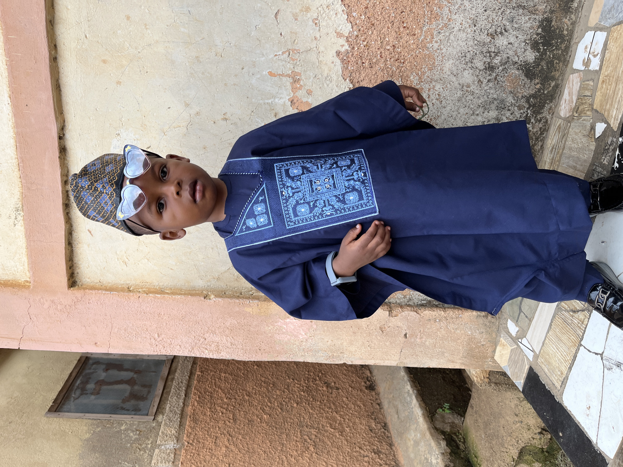 A young boy is standing outdoors, wearing a traditional blue garment with intricate embroidery and a matching patterned cap. He is also wearing heart-shaped sunglasses. He is standing on a tiled step next to a building. The building has a textured, multi-toned wall in shades of beige, brown, and pink. A small, screened window is visible near the bottom of the wall. The scene appears to be in Jos, Nigeria, based on the style of dress and the environment.