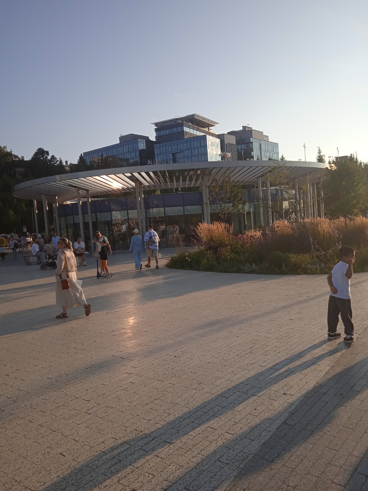 The image depicts an outdoor scene in Kazan, Russia, likely near the "Kazan Family Center" (also known as the "Chasha" or "Cup") due to the distinctive modern architecture of the building in the background and the presence of a circular, colonnaded structure in the foreground.
In the foreground, a boy in a white t-shirt and gray pants stands on a paved walkway, facing to his right. His right hand is raised to his face, and his shadow is cast long across the walkway. To the left of the walkway, a woman in a light-colored dress and sandals walks away from the camera, carrying a brown purse. Another woman on a scooter rides past her, and several other people are seated on benches or walking in the background, near the circular structure.
The circular structure has a metal roof supported by numerous thin columns. Large glass windows or doors are visible behind the columns. Behind the circular structure, a multi-story modern building with glass facades rises. Trees and other vegetation are present in the middle ground and background. The sky is clear and blue, indicating fair weather, and the sun is low in the sky, casting long shadows. The overall impression is of a public park or plaza during the late afternoon or early evening.