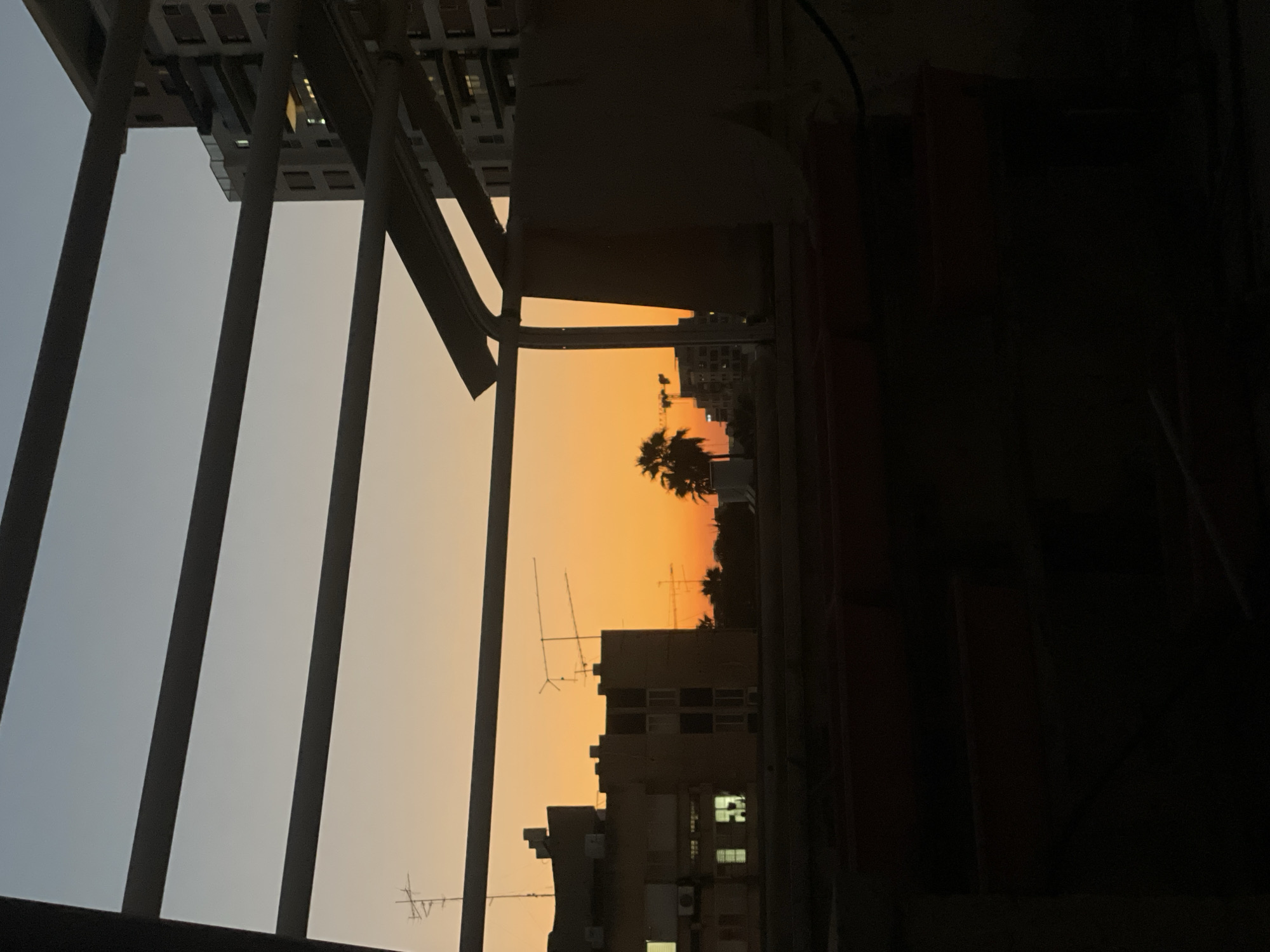 The image captures a sunset over the city of Petah Tikva, Israel, viewed from an elevated perspective. The sky transitions from a pale blue at the top to a vibrant orange near the horizon, indicating the time of day. Silhouetted against the sky are various urban elements. In the foreground, dark, indistinct structures, possibly seating or planters, are visible.
Further into the scene, buildings are discernible, with some windows illuminated. Rooftop antennas are prominent on several structures. A palm tree is silhouetted to the left of the frame, suggesting a warm climate. In the background, construction cranes are faintly visible, hinting at ongoing development within the city. The overall scene is framed by dark, horizontal and angled beams, likely part of a balcony or rooftop structure, which add a sense of depth and enclosure. The lighting is low due to the sunset, creating strong contrasts between illuminated areas and deep shadows.