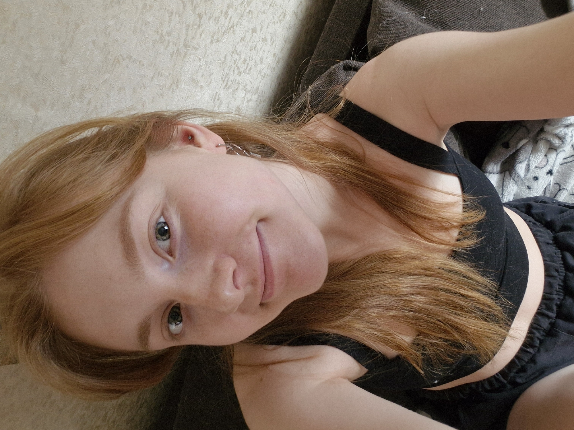 A young, light-skinned person with auburn hair and light-colored eyes is positioned in a selfie-style shot. The individual is lying down, with their head tilted towards the camera. They are wearing a black sports bra and black shorts. The background features a light-colored wall and a dark-colored couch or bedding. The location is unknown, there is no information indicating this photo was taken in Yekaterinburg, Russia.