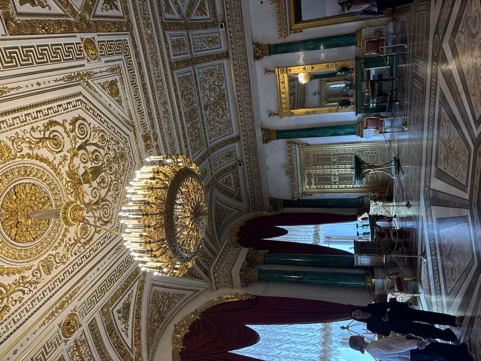 The image depicts an ornate interior space, likely a room within a historic building in Saint Petersburg, Russia. The ceiling is highly decorated with gilded ornaments and geometric patterns surrounding a large, multi-tiered chandelier. The walls feature marble columns and intricate details. Red drapery is visible near window areas. The floor is patterned with inlaid stonework. Two people are visible in the foreground, one wearing a light shirt and another dressed in dark clothing, standing close to the windows.