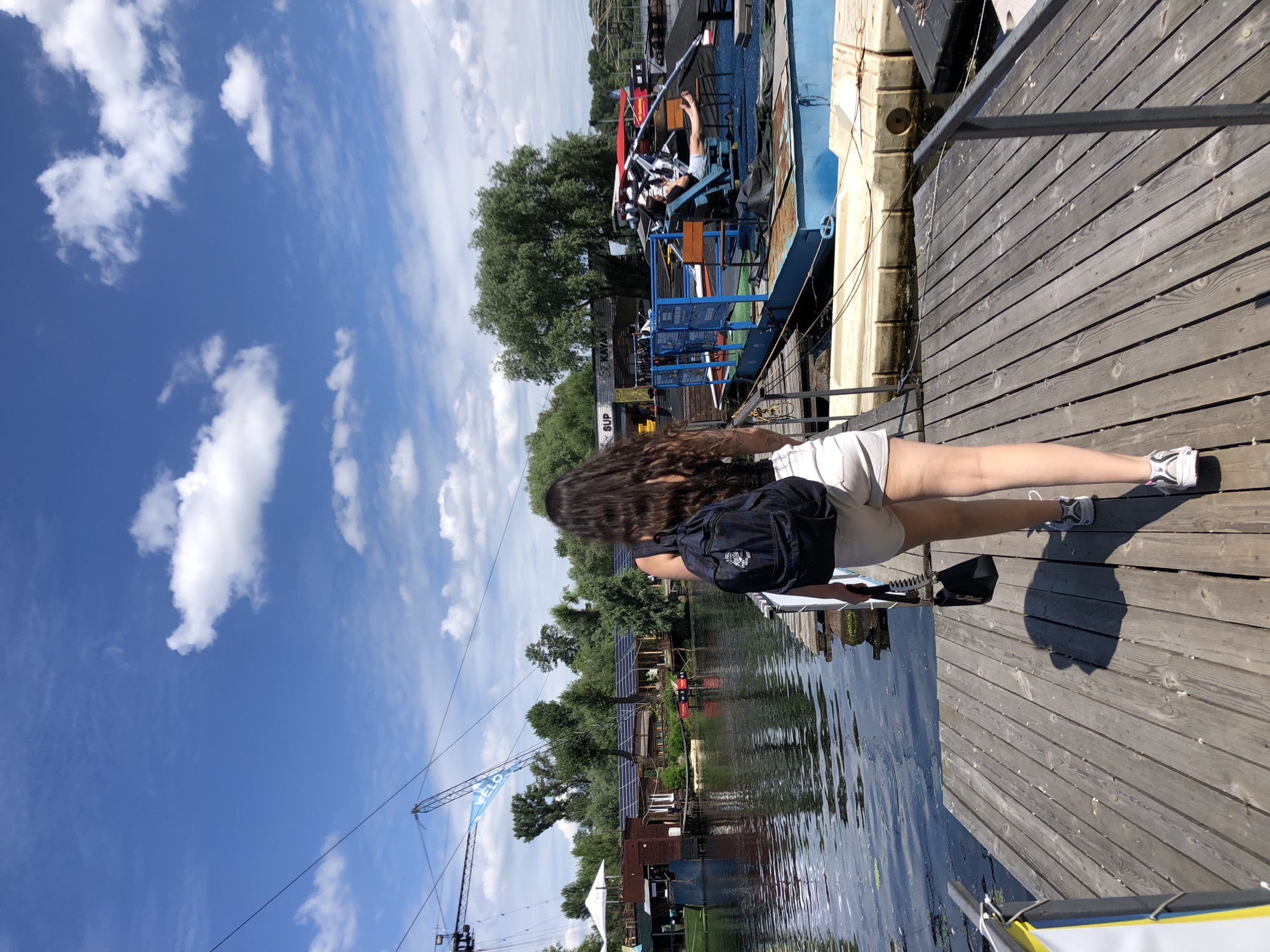 A young woman with long, dark, wavy hair walks away from the camera on a wooden pier. She is wearing a black tank top, light-colored shorts, and athletic shoes. She has a dark backpack on her back and a small black purse in her right hand. The pier is next to a body of water with several boats and structures on the opposite bank. In the background, there are trees, buildings with solar panels, and a wakeboarding cable system. The sky is blue with scattered white clouds. The scene appears to be an outdoor recreational area on a sunny day.