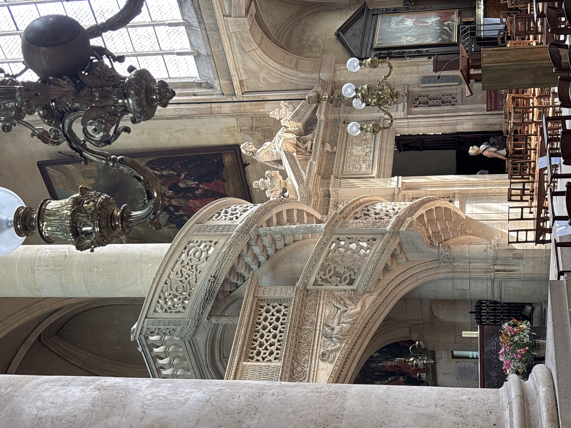 This is an interior photograph of a grand stone staircase within a Parisian church. The elaborate spiral staircase, carved from light-colored stone, features intricate lattice work and sculpted reliefs. A large, ornate brass chandelier hangs from the ceiling, casting a warm glow on the scene. In the background, a large framed painting hangs on the wall, depicting a religious scene. Sunlight streams through a large arched window with multiple panes, illuminating dust motes in the air. The environment appears to be a place of worship, with rows of wooden chairs arranged in the foreground, suggesting it is a church or cathedral. A person is visible walking through an open doorway in the mid-ground. The architecture is detailed and appears historic, indicative of a significant Parisian landmark.