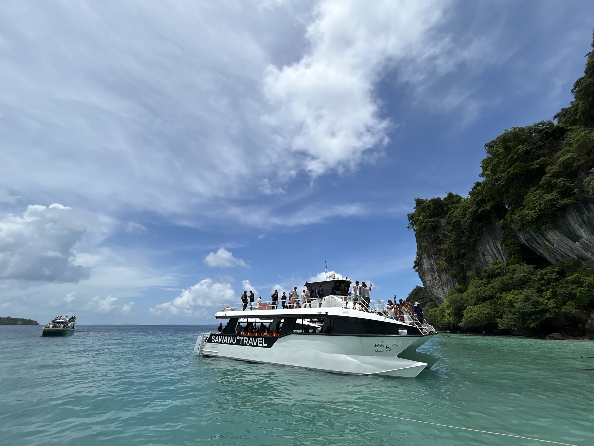 The image depicts a bright, sunny day with a clear blue sky dotted with white clouds. The scene is set on the turquoise waters of the sea, likely near Ton Sai, Thailand, given the characteristic karst limestone cliffs covered in lush green vegetation. In the foreground, a white catamaran named "SAWANU TRAVEL" is prominently featured. The boat is carrying numerous passengers, with several individuals standing on the upper deck and more seated inside the enclosed lower cabin. The boat is moving, indicated by the slight ripple in the water around its hull. Further in the background, another, smaller boat is visible, also with people on board. The overall impression is one of a tourist excursion or a ferry service operating in a scenic coastal area.