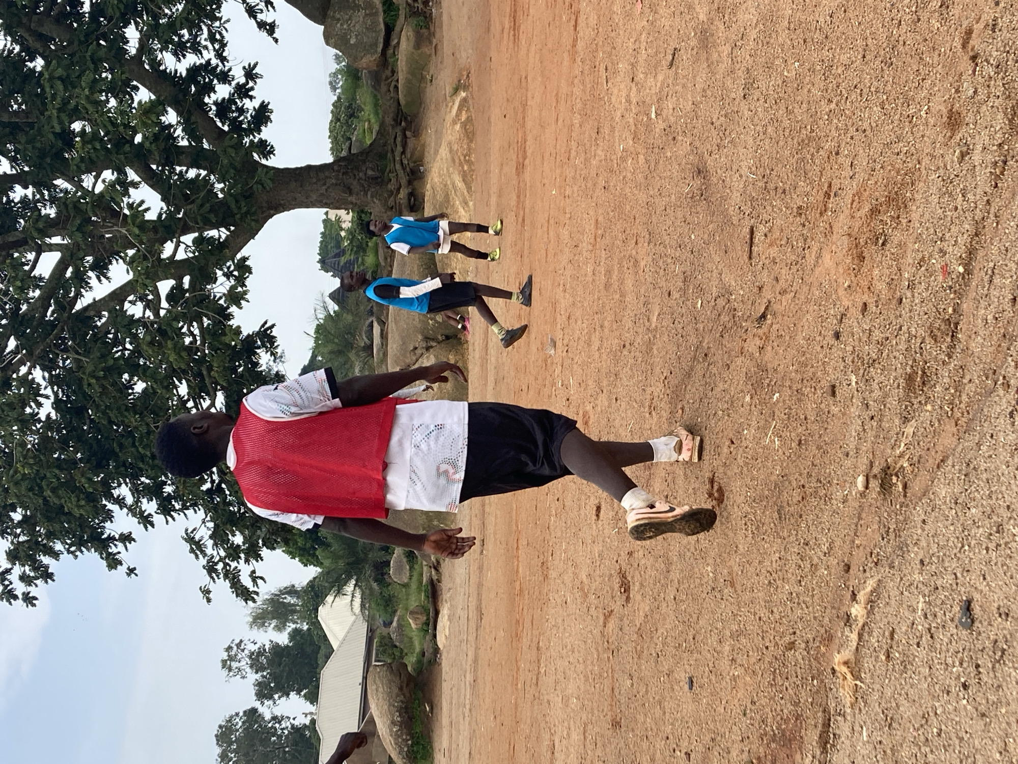 A group of young men are playing soccer on a dirt field in front of a large tree in Jos, Nigeria. The man in the foreground is wearing a white t-shirt with a red vest, black shorts, and pink sneakers. He is walking away from the camera. Two other young men are in the background, also wearing blue vests and shorts. The field is dry and dusty, with scattered rocks and vegetation. The sky is overcast.