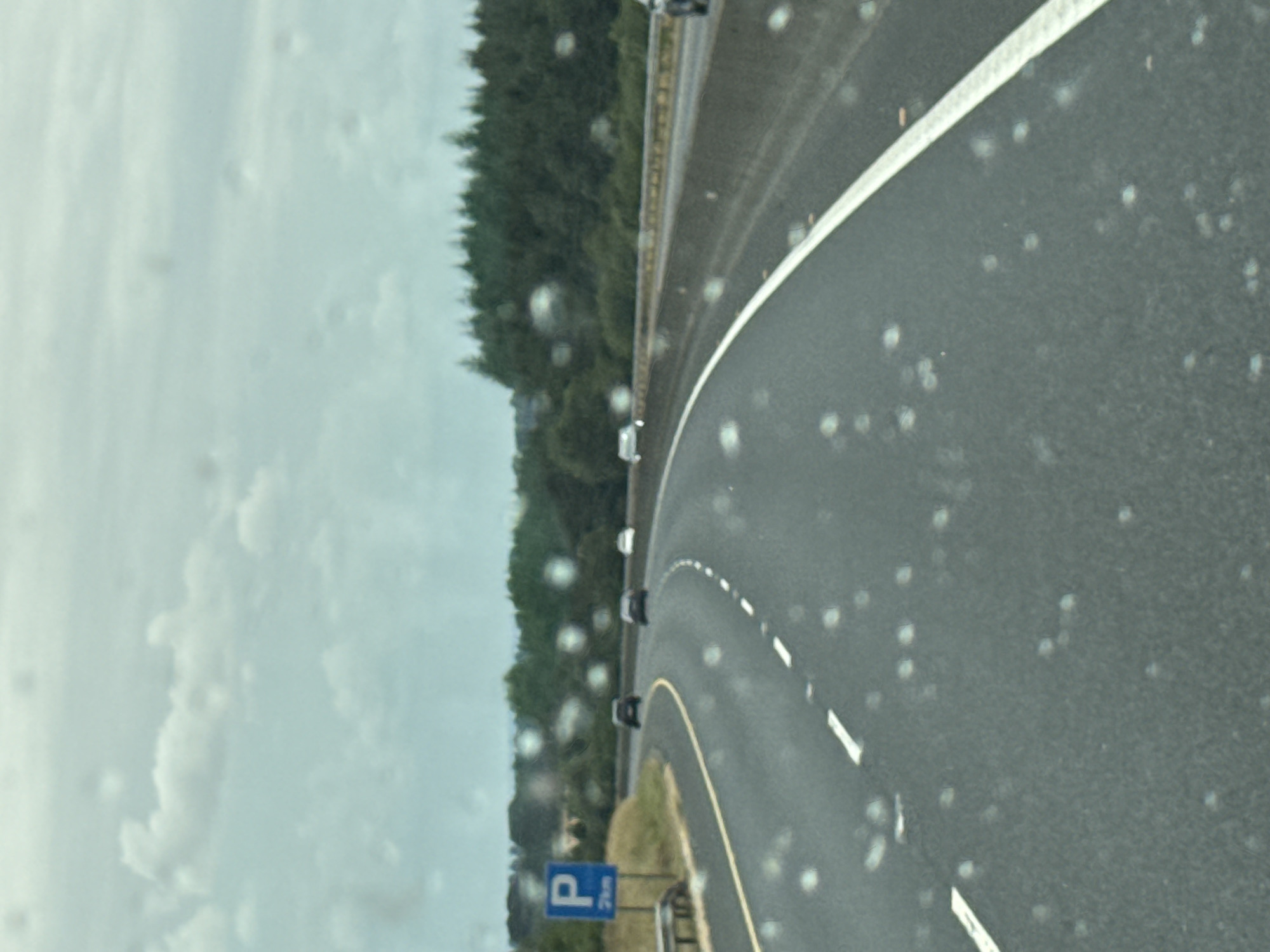 The image depicts a view from a vehicle traveling on a highway, likely the M7 motorway, near Colbinstown, Ireland. The road curves to the right, with two lanes visible in the direction of travel. The road surface is asphalt, marked with white dashed lines. A blue sign with a white "P" indicates a parking area is 2 kilometers away, positioned on the left side of the road. On the left shoulder of the road, there is a wooden barrier. The right side of the road is bordered by a concrete barrier. In the distance, there is a dense line of evergreen trees forming a forest. The sky is overcast with visible clouds, and there are numerous small water droplets on the windshield, indicating rain or a wet surface. Several cars are visible on the highway, appearing as dark shapes in the distance.