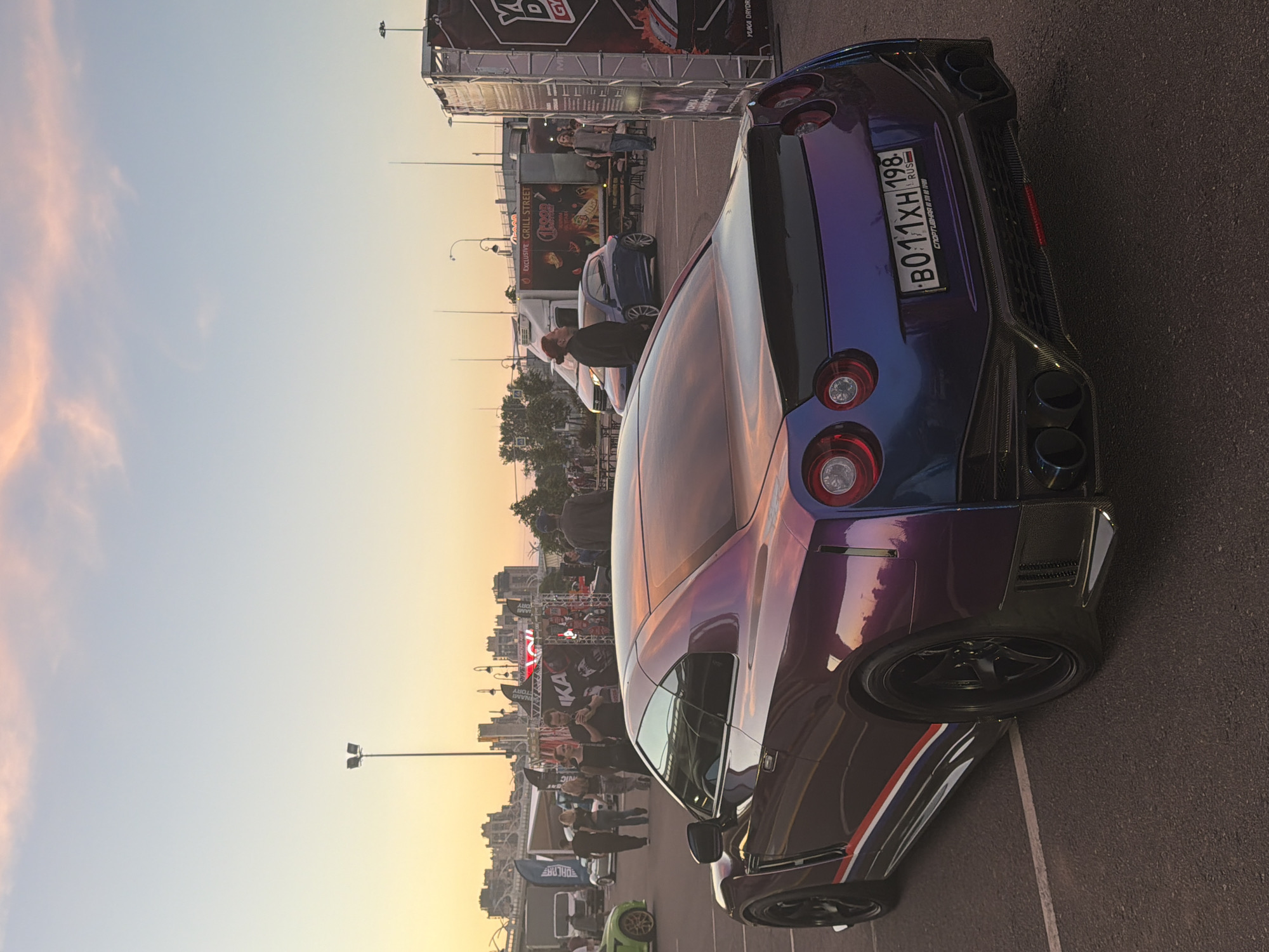 A modified Nissan GT-R with a iridescent purple and blue wrap is parked on asphalt in an outdoor lot, likely at a car show or event, as indicated by the tents and crowd in the background. The car is viewed from the rear, three-quarter angle, showcasing its custom body kit, quad exhaust pipes, and racing-style wheels. A Russian license plate is visible on the rear bumper. The sky above is a soft gradient of orange and pink hues, typical of sunset. Several individuals are present in the background, some walking and others standing near tents and vehicles. A food truck is also visible in the mid-ground. The overall atmosphere suggests an evening event related to automobiles.
