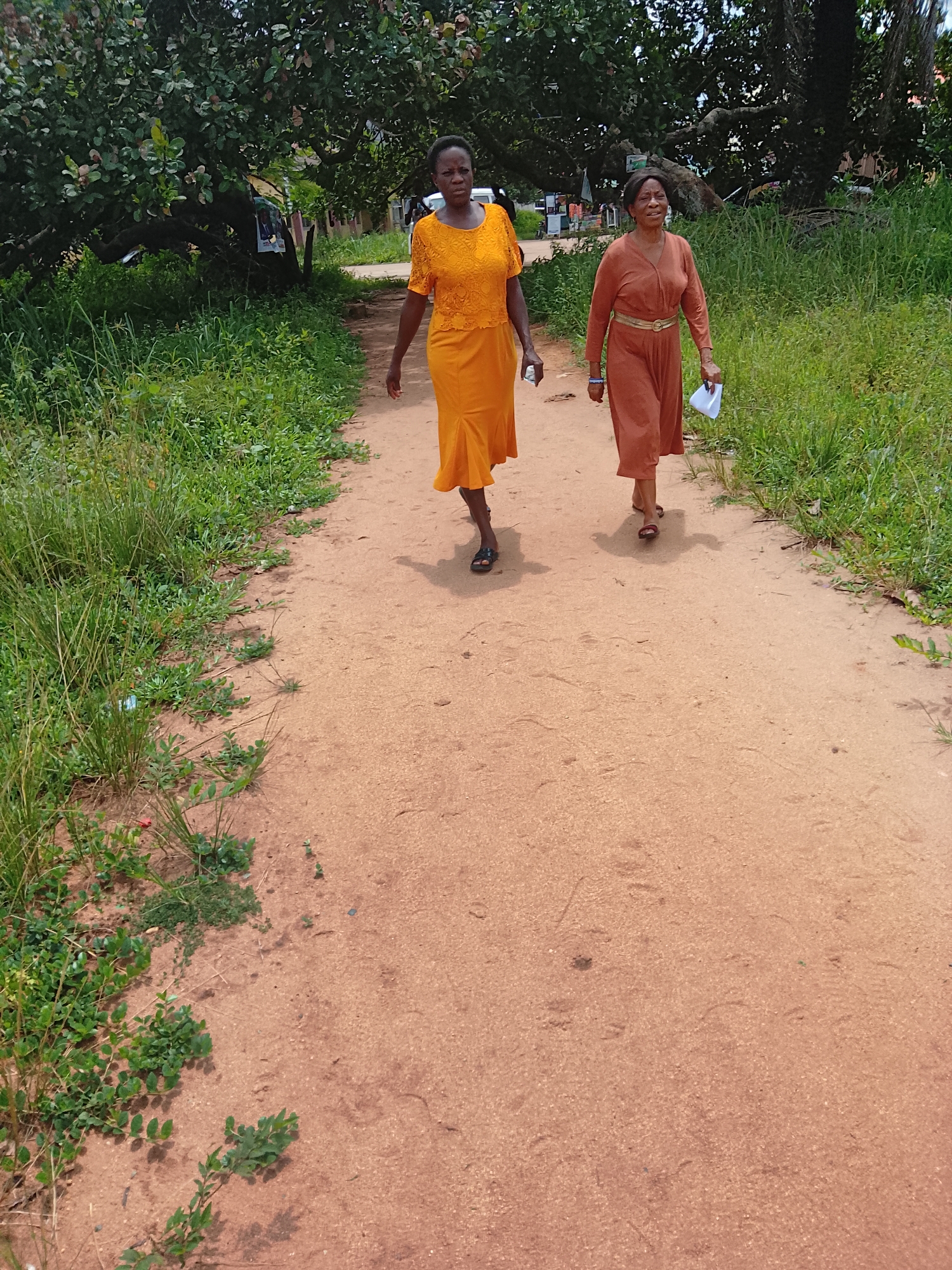 In Ihiagwa, Nigeria, two women are walking on a dirt path. The woman on the left wears a bright orange, lace-patterned dress and dark sandals. The woman on the right wears a rust-colored dress with a gold belt and red sandals, carrying a white piece of paper. They are walking side-by-side on a reddish-brown dirt path that is bordered by lush green grass and vegetation on the left. In the background, tall trees with dense foliage provide shade, and hints of buildings and a few parked vehicles are visible. The sky is overcast, suggesting a shaded environment. The overall scene depicts a casual outdoor stroll in a semi-rural or suburban area.