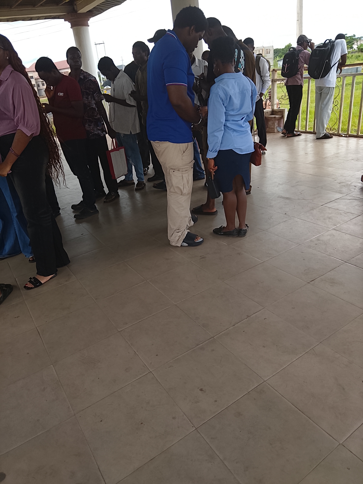 An outdoor, tiled ground level scene in Ihiagwa, Nigeria shows a group of approximately ten people standing in a line. The people are of varying genders and ethnicities, and are dressed in casual clothing. Some individuals are looking down at their phones, while others appear to be conversing or observing their surroundings. The group is positioned under a covered walkway or veranda, with a railing and grassy area visible in the background. The overall impression is that of people waiting or gathered in a public space.