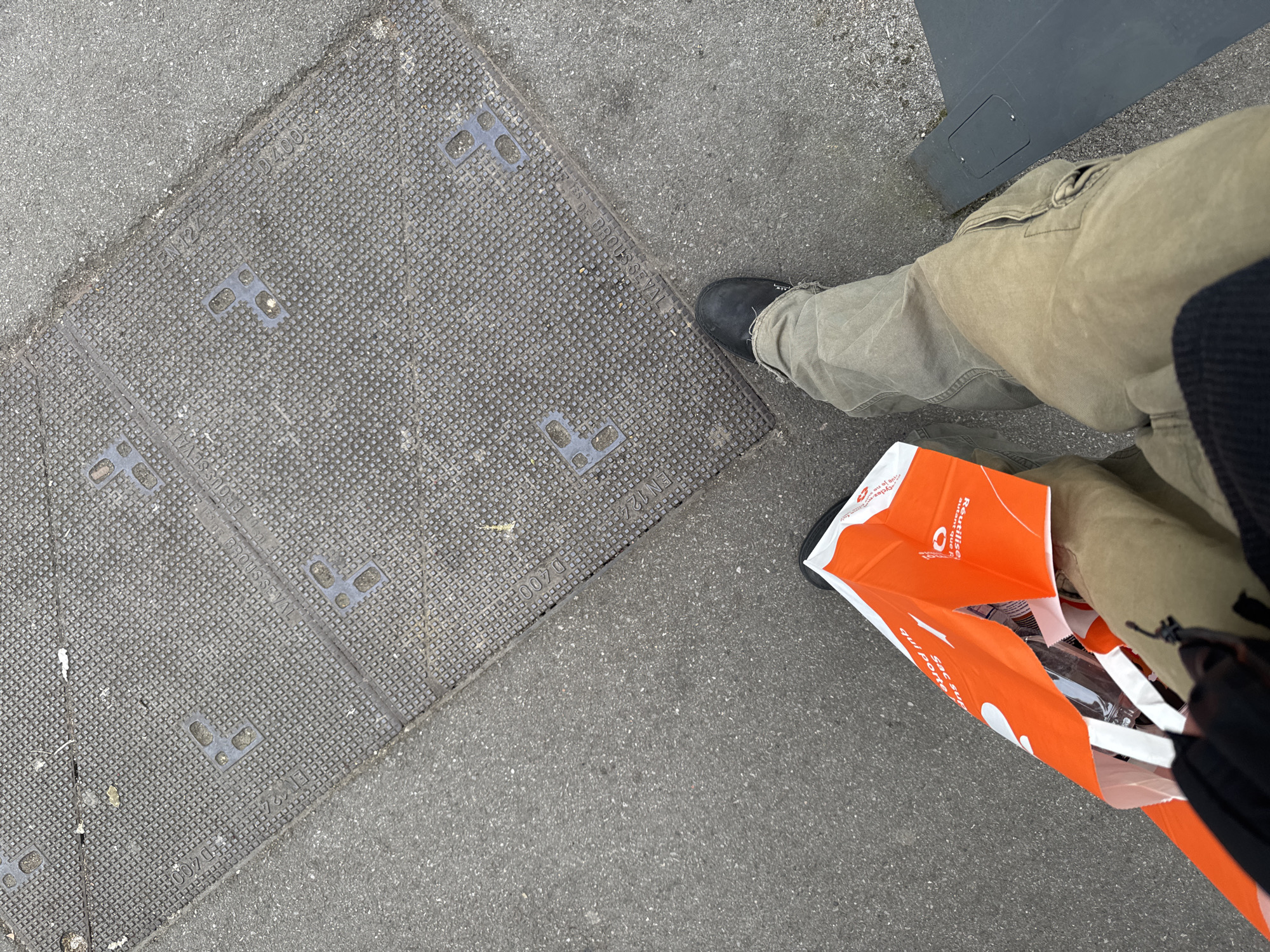 The image captures a street scene, focusing on a square utility access cover on a paved surface. The cover features a textured, dotted surface and multiple square indentations. In the upper right quadrant, a person wearing khaki trousers and dark shoes is partially visible. An orange shopping bag with white text is prominently displayed near the person's legs. The background consists of the paved surface and a dark gray element in the upper right corner. OCR identifies the names "BROUSSEVAL" and codes "EN124", "D400" and text that relates to reusable shopping bags. The mention of "Brousseval" is linked to the city of Les Ulis, France, suggesting this is the location.