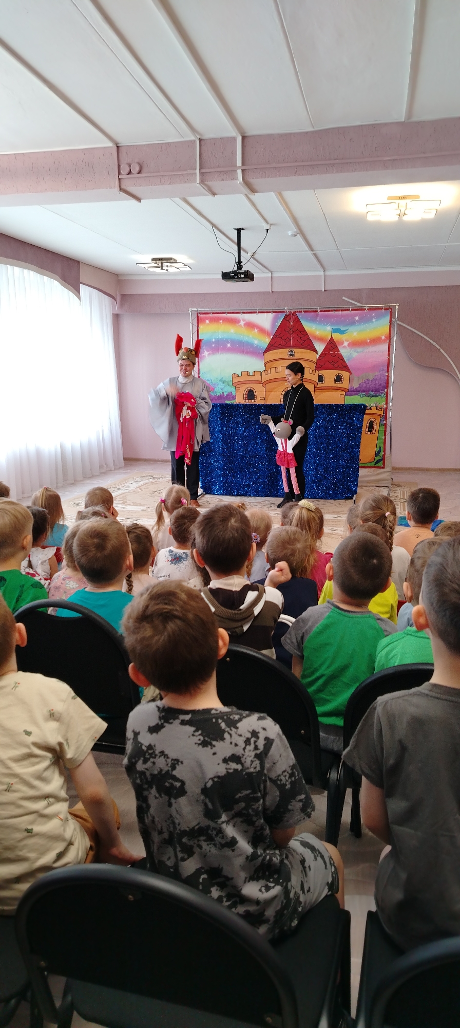 A group of children are seated on black folding chairs watching a puppet show at an indoor event in Chaikovsky, Russia. The setting appears to be a brightly lit room with a white ceiling, potentially a classroom or community hall. Two individuals are performing the puppet show on a stage. The background features a banner with a castle and rainbow design. One performer is dressed in a light-colored robe with decorative headwear, while the other is in a black outfit operating a mouse puppet. The children are mainly facing away from the camera, showing their backs and the backs of their heads.