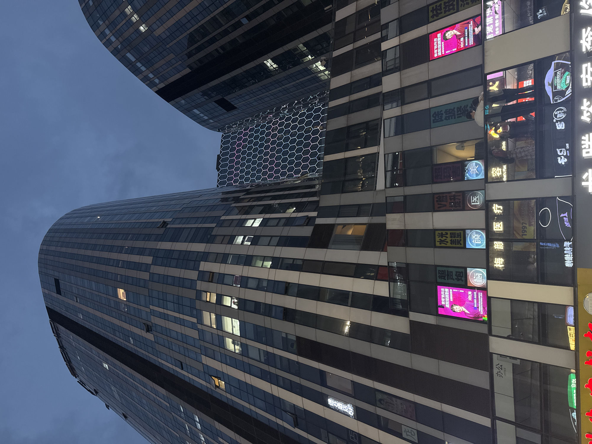 The image depicts a nighttime view of modern high-rise buildings in the Chaoyang District of China. The buildings feature contemporary architecture with extensive glass facades. Some windows are lit, suggesting occupancy. Multiple illuminated signs and advertisements are visible on the lower levels of the buildings, indicating commercial activity. One building in the background has a distinctive hexagonal pattern illuminated with colorful lights. The sky is dark, indicating it is either dusk or night. A few people are visible, one pair embracing on a balcony area. The overall scene conveys a sense of urban nighttime vibrancy.