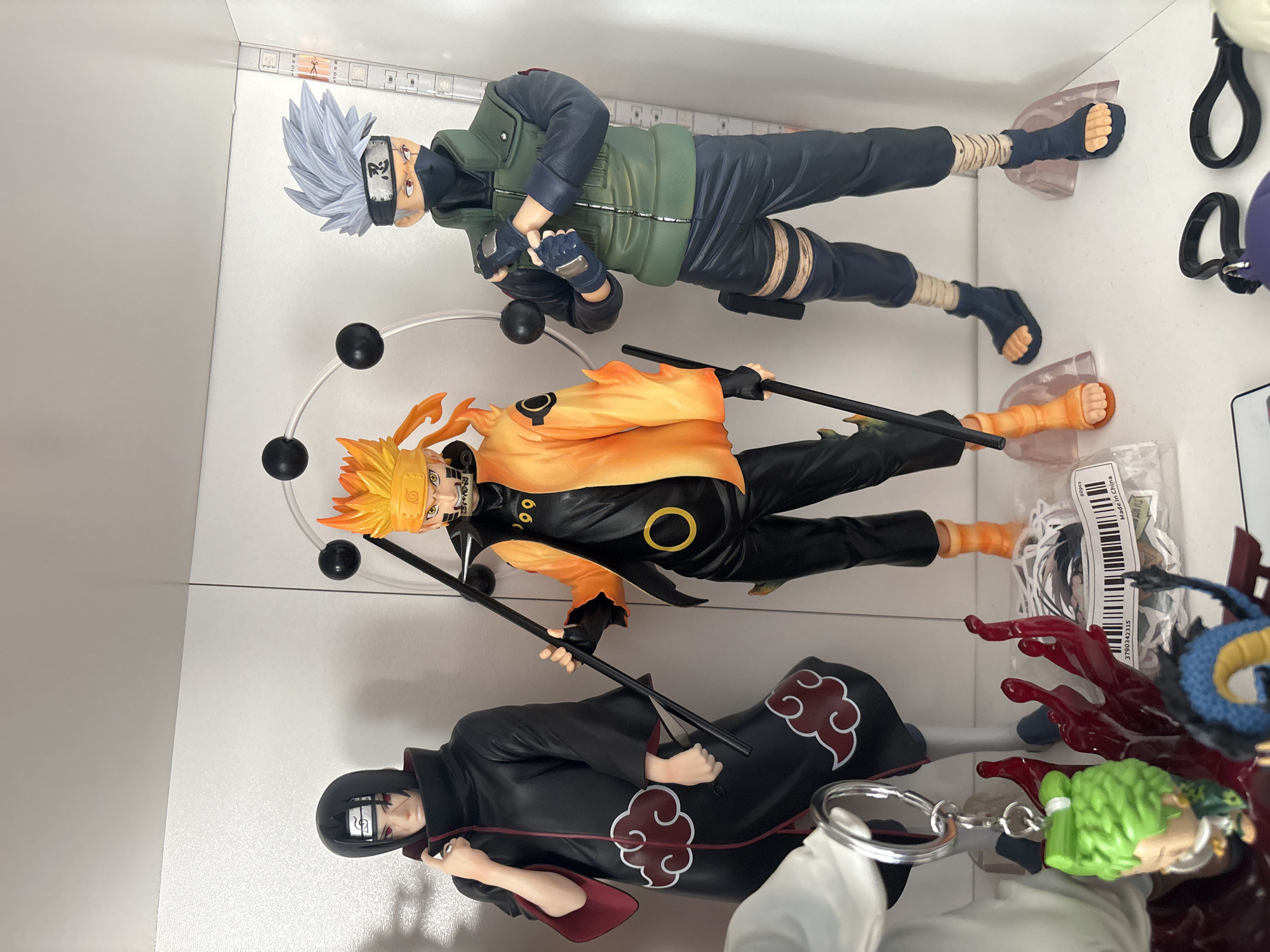 The image shows a display shelf with three Naruto action figures. From top to bottom, the figures are Kakashi Hatake, Naruto Uzumaki (Six Paths Sage Mode), and Itachi Uchiha. Other items include keychains, but the action figure details are difficult to determine. The display is in a brightly lit, enclosed space. The exact location in Plovdiv, Bulgaria, cannot be determined from the image.
