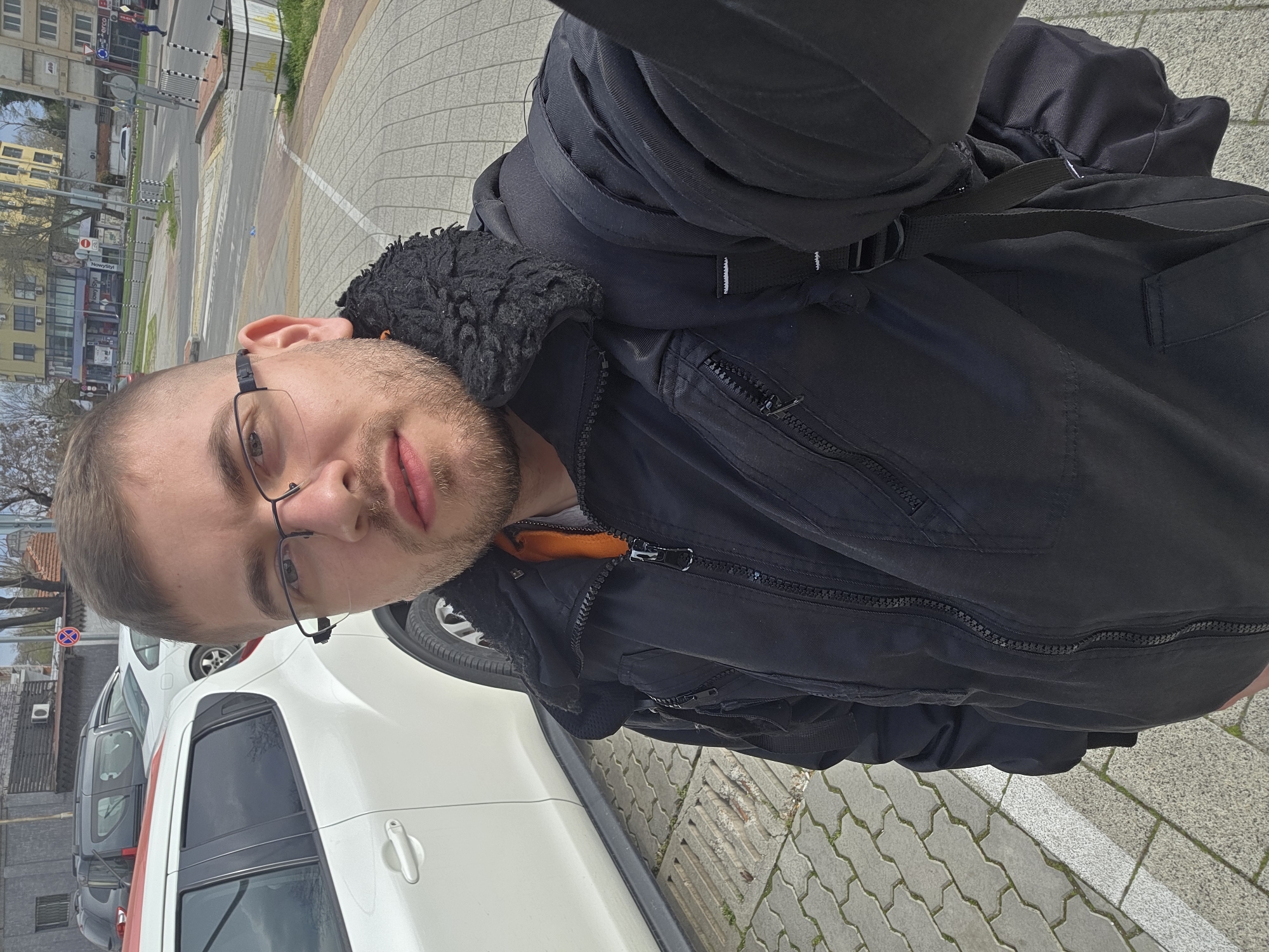 A man with short brown hair, a beard, and glasses is standing on a sidewalk in Plovdiv, Bulgaria. He is wearing a black jacket with an orange liner and a black backpack. He is facing the camera with a neutral expression. To his left is a white car. The sidewalk is made of gray paving stones. In the background, there are buildings, trees, cars, and street signs, suggesting a typical urban environment.