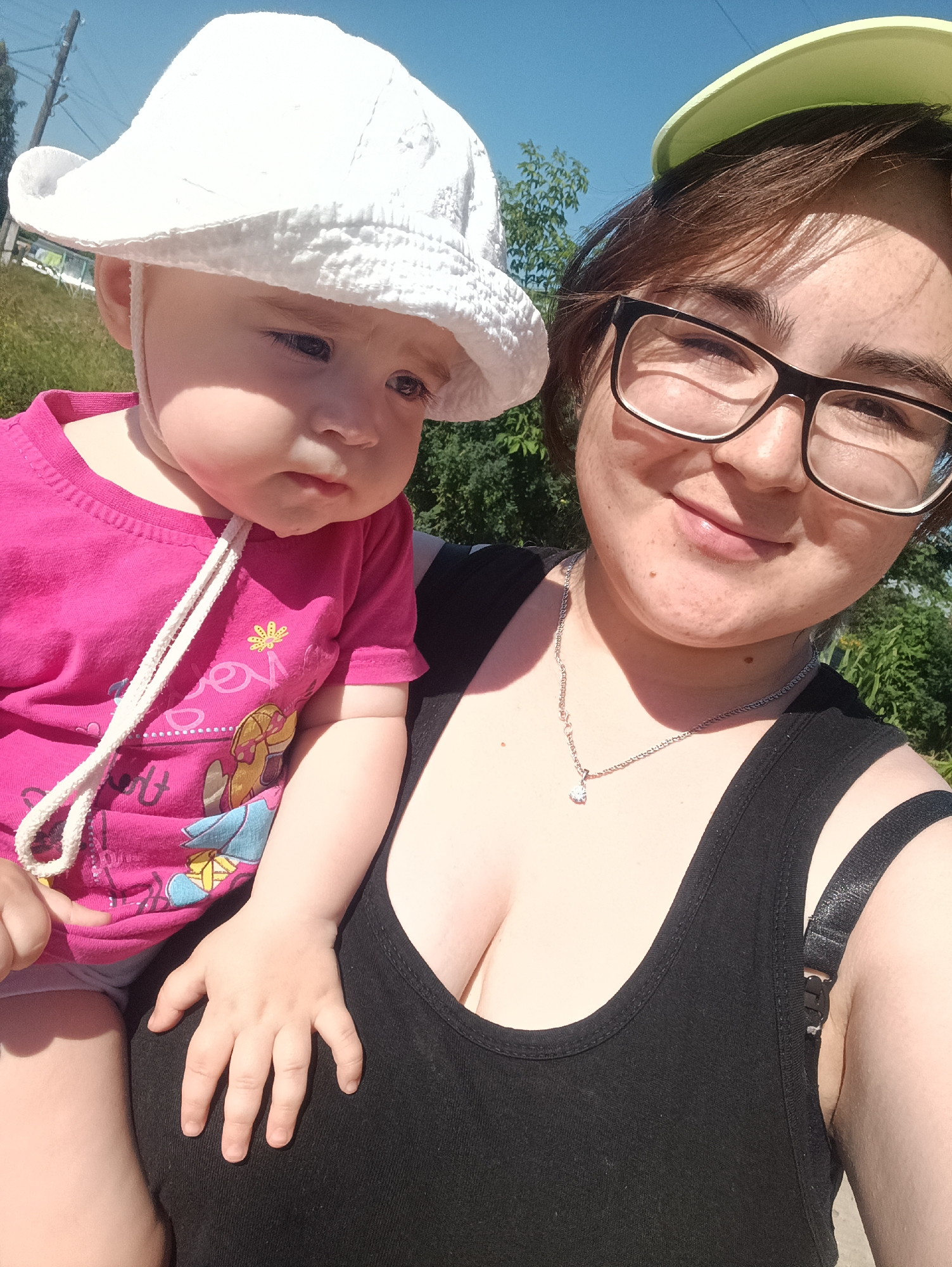 This image is a selfie taken outdoors on a sunny day. A woman is in the foreground, wearing a black tank top and glasses. She is holding a baby who is wearing a white sun hat and a pink t-shirt. The background shows trees and a blue sky. The approximate location of this image is in the city of Posad, Russia, indicated by the Cyrillic text on the pink baby's t-shirt which says "ПОСАД".