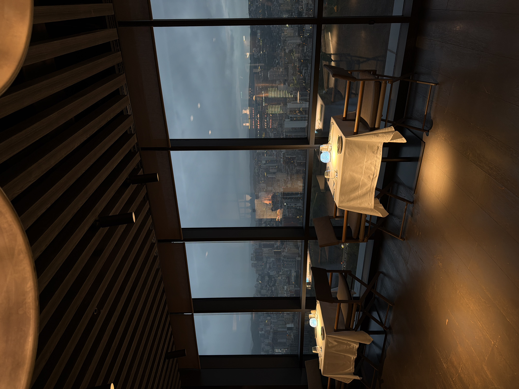 The image displays an interior view of a high-rise restaurant or bar at dusk or early night, looking out over a city skyline. The original image is rotated 90 degrees clockwise.
The scene features a sophisticated dining area with large, floor-to-ceiling windows revealing a panoramic view of a city. The sky outside is a soft, dark gray-blue, indicating either a cloudy evening or early night, with the distant city lights beginning to illuminate the urban sprawl. Several buildings below show illuminated windows, and faint reflections of the city are visible on the windowpanes.
Inside, the lighting is warm and dim. The ceiling on the left is adorned with dark wooden slats arranged diagonally, fitted with subtle spotlights that cast a gentle glow. A large, circular light fixture or decorative element is partially visible in the upper left corner.
Along the window, several dining tables are set with crisp white tablecloths. On one table, glasses, plates, and a distinctive blue, patterned table lamp emitting a soft, ambient light are visible. Elegant wooden chairs with light-colored upholstery are placed around the tables. The floor is made of dark wood, reflecting the warm artificial lights from the ceiling and the table lamps.
No people are present in the image, suggesting the restaurant might be awaiting patrons or is captured after closing. The overall atmosphere is quiet, luxurious, and evokes a sense of calm and anticipation. There is no visible text in the image.