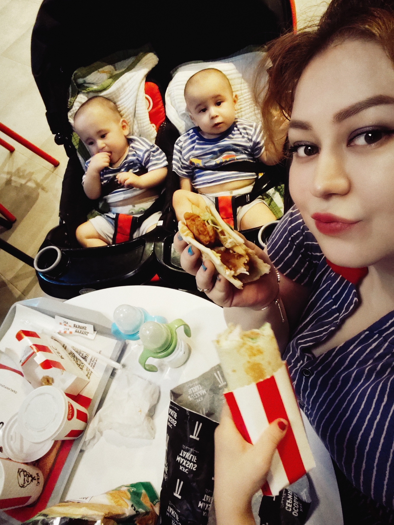In Fergana City, Uzbekistan, a woman is taking a selfie with two babies in a stroller, holding a half-eaten wrap. The babies, dressed in matching blue and white striped outfits, are seated in a black stroller. One baby is looking down and has its fingers in its mouth, while the other baby is looking towards the camera. The woman, with reddish-brown hair and dark eye makeup, is wearing a blue and white striped shirt. In the foreground, a white table holds a KFC tray with various food items, including a wrap, cups, and napkins, as well as two baby bottles. The background shows red chair legs and a beige tiled floor. The overall scene suggests a casual outing, possibly at a fast-food restaurant.