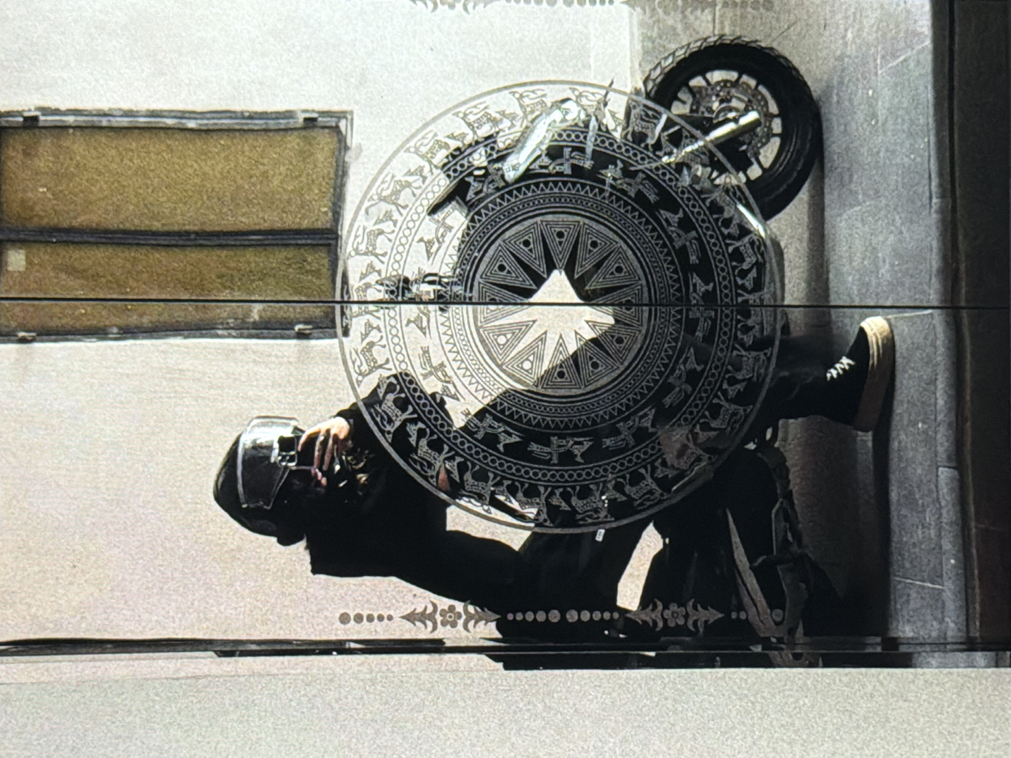 A man wearing a black motorcycle helmet, black clothing, and black shoes, lies on a tiled ground next to a grey concrete wall in Đà Nẵng, Vietnam. A motorcycle is partially visible. A large bronze drum is positioned above him. A rectangular window-like structure is located high on the wall. The drum has intricate patterns, and the tiled ground has decorative inlays.