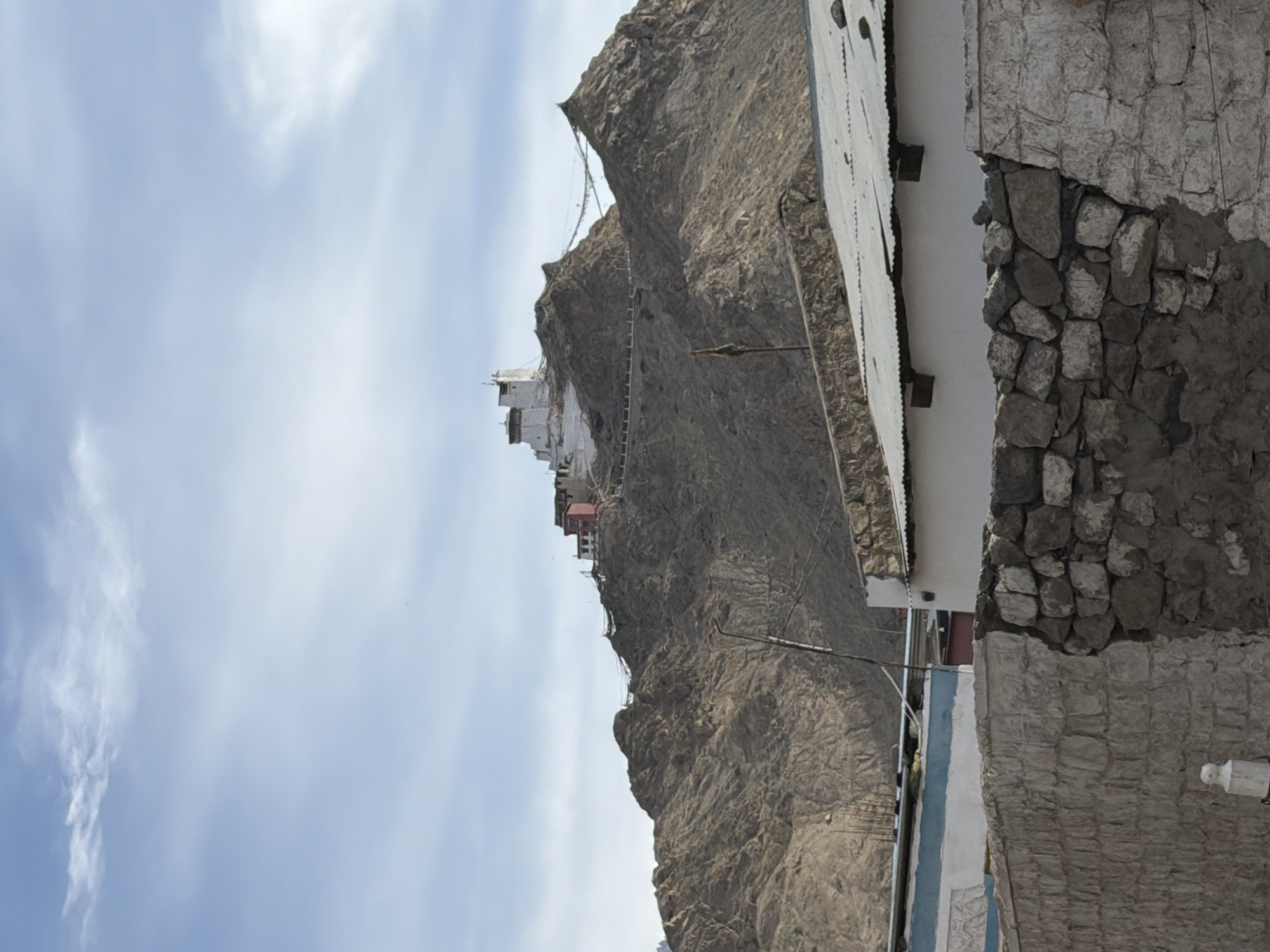 The image depicts the Tsemo Monastery in Leh, India, situated atop a rocky mountain. The monastery's white structure is prominent against the vast, cloudy sky. In the foreground, there are some low-rise buildings with corrugated tin roofs and stone walls, suggesting a residential or market area. Prayer flags are strung between the peaks, a common sight in Buddhist regions. The overall scene is one of a mountain settlement with religious significance.