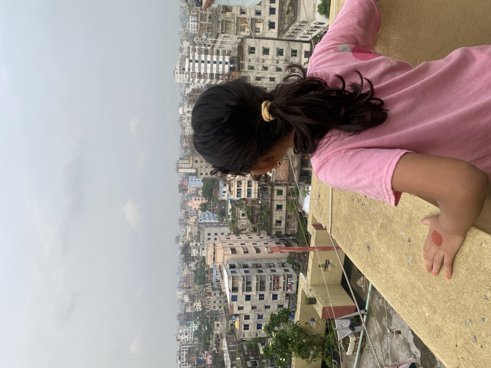 A young girl with dark hair tied in a ponytail, wearing a pink t-shirt, stands on a rooftop overlooking a dense urban landscape of multi-story buildings under a cloudy sky. Her left hand, with manicured orange fingernails, rests on the yellow railing of the rooftop, displaying a circular reddish design on her wrist. The buildings in the background appear to be a mix of residential and commercial structures, with varying architectural styles and colors, indicative of a bustling city. Laundry hangs on clotheslines between some of the buildings, and vegetation is visible on some rooftops. The overall scene suggests a typical day in a densely populated area of Dhaka, Bangladesh, characterized by its extensive urban sprawl.