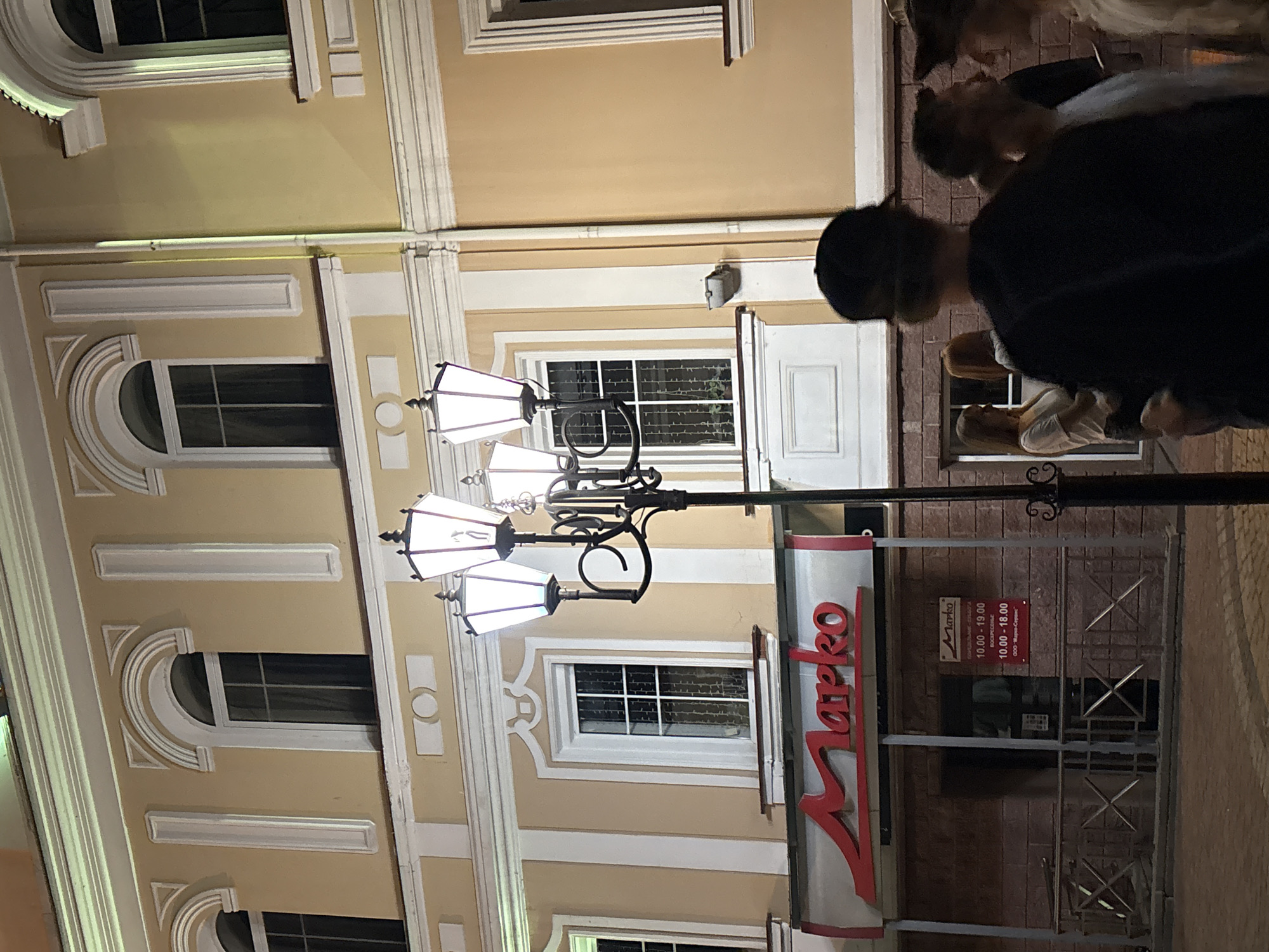 The image shows a street scene at night in Vitsebsk, Belarus, with a building facade illuminated by streetlights. The building has several arched windows with white trim and is painted a light yellow color. A prominent red "Marko" sign is visible above the entrance to a shop. In the foreground, a black lamppost with multiple lights stands tall. Several people are present in the scene, some silhouetted against the building and others in conversation. The ground appears to be paved.