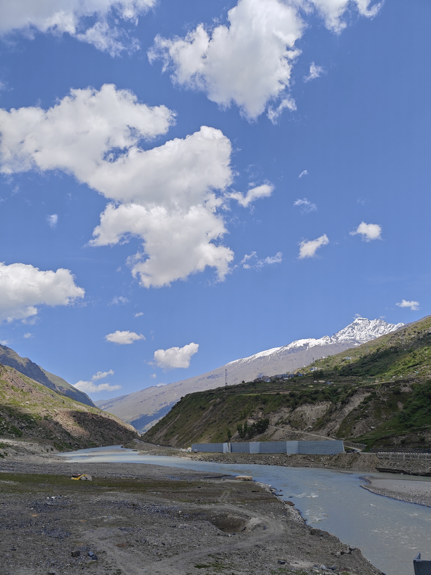 This is a wide shot of a river flowing through a valley. The river is wide and shallow, with a light blue color. The valley is steep and rocky, with green vegetation on the slopes. In the background, there are mountains, some of which are covered in snow. The sky is blue with some white clouds. There is a yellow excavator on the left side of the river. There are also some buildings on the hillside in the distance. The scene appears to be from a sunny day.