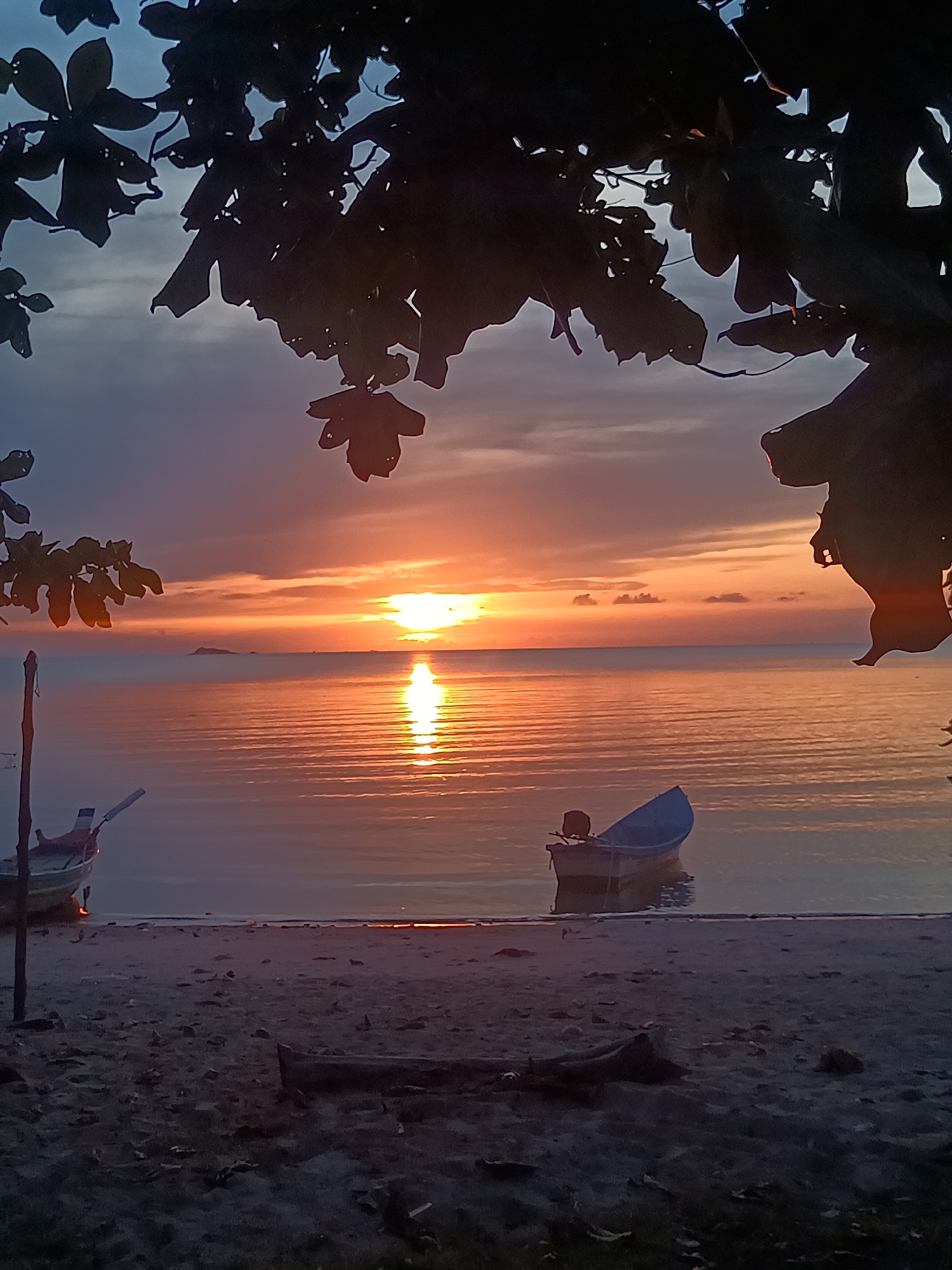 This is an eye-level, outdoor shot of a serene beach at sunset, likely in Baan Wok Tum, Thailand. Silhouetted tree branches and leaves partially frame the top of the image. Below, the horizon shows a vibrant sunset with an orange glow, reflected in the calm water. Two boats are visible: one near the left edge and one in the center, both appearing to be resting close to the shore. The beach in the foreground is sandy and dark, with some driftwood or debris scattered on it. The overall impression is one of tranquility.